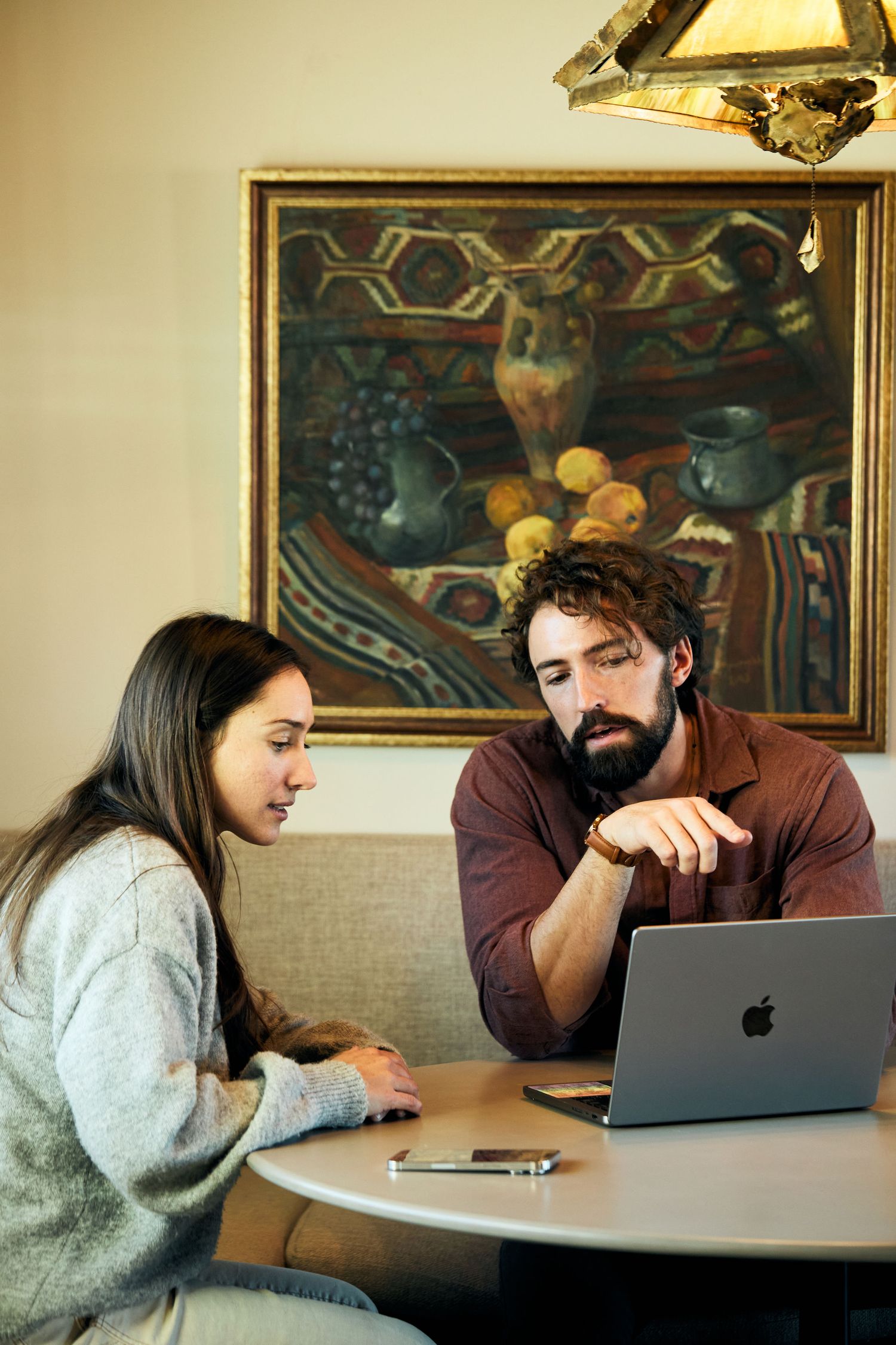 Two people have a discussion while sitting at a round table with a MacBook laptop and decorative artwork on the wall.