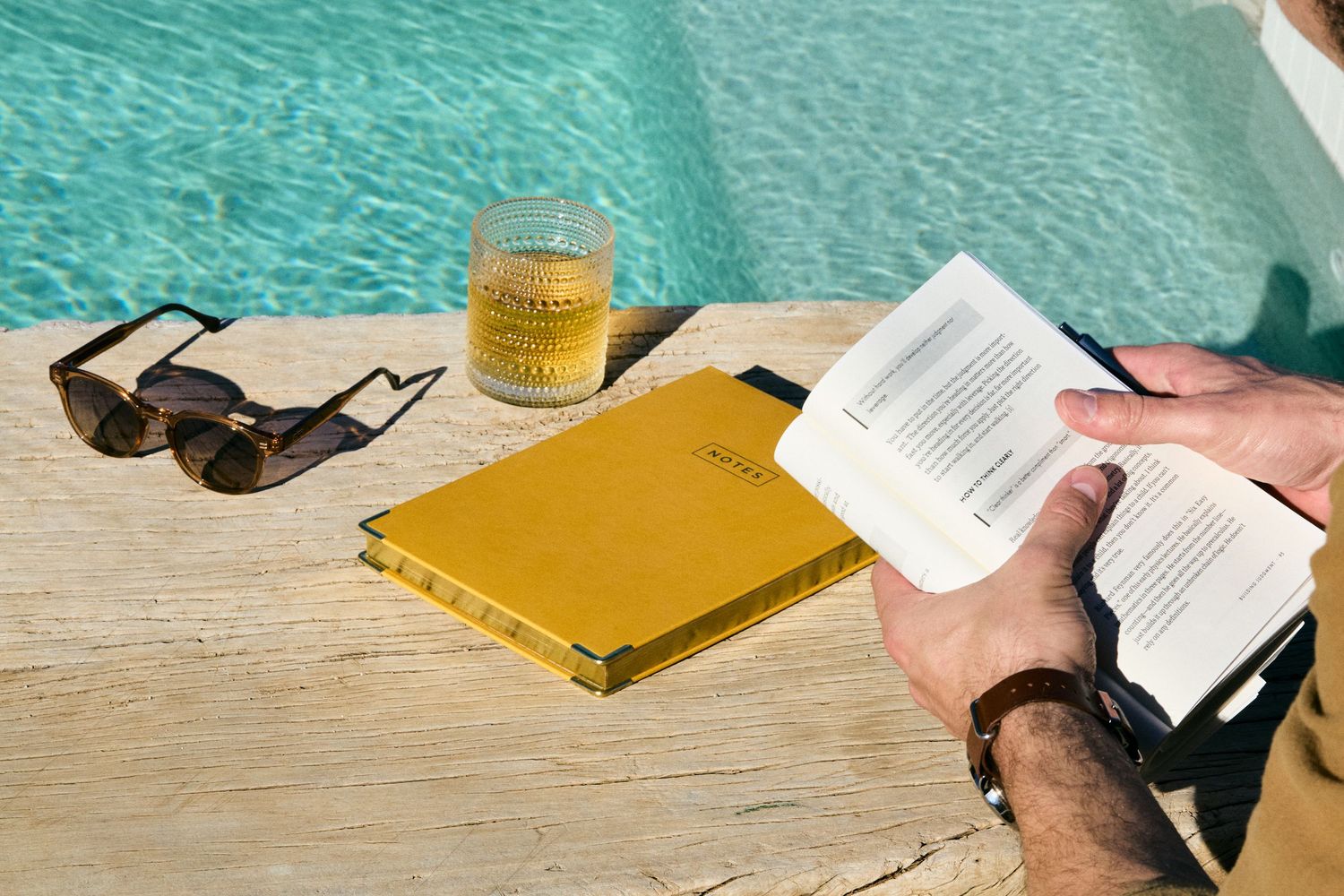 A person reads a book while relaxing next to a turquoise swimming pool on a sunny day.