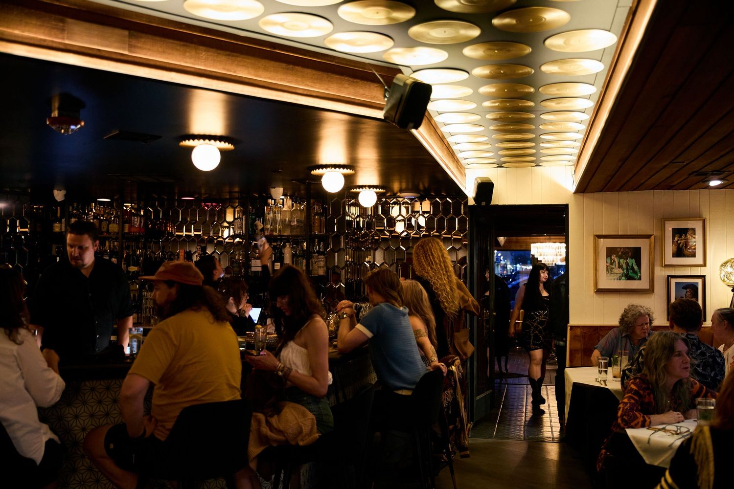 Dimly lit bar interior with patrons seated at counter under warm lighting and exposed ceiling beams.