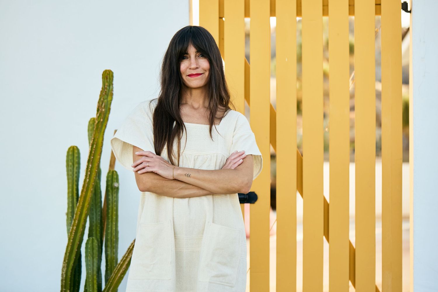 Someone in a white linen dress stands with crossed arms next to a tall cactus against a yellow slatted wall.