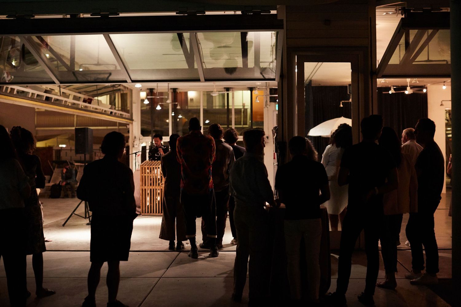 People gather outside an illuminated building entrance at night, creating silhouettes against the warm interior lighting.