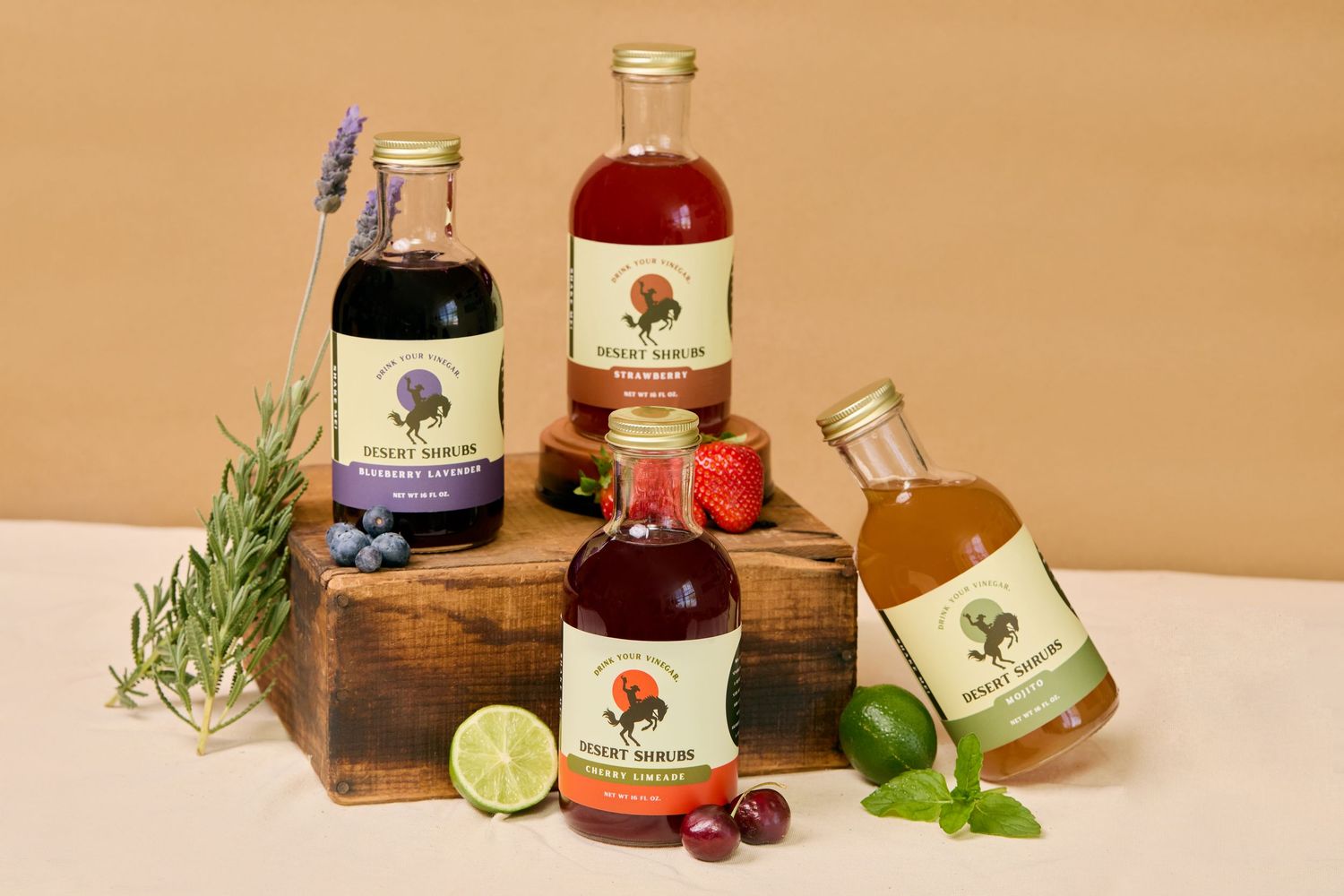 Four bottles of fruit syrups surrounded by fresh herbs and fruits on a wooden block against a beige background.