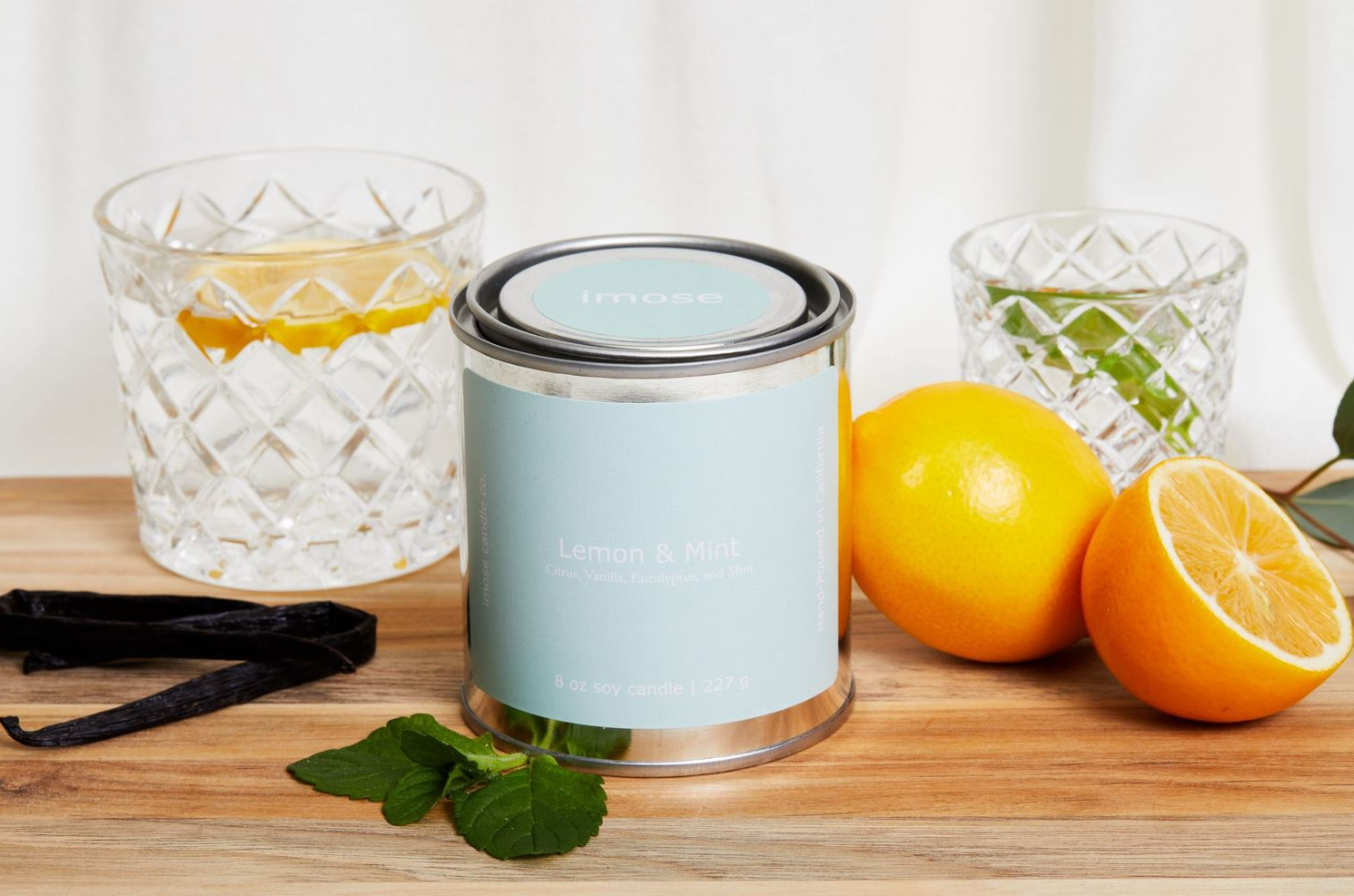 Light blue candle tin displayed with citrus fruits and crystal glasses on wooden surface.