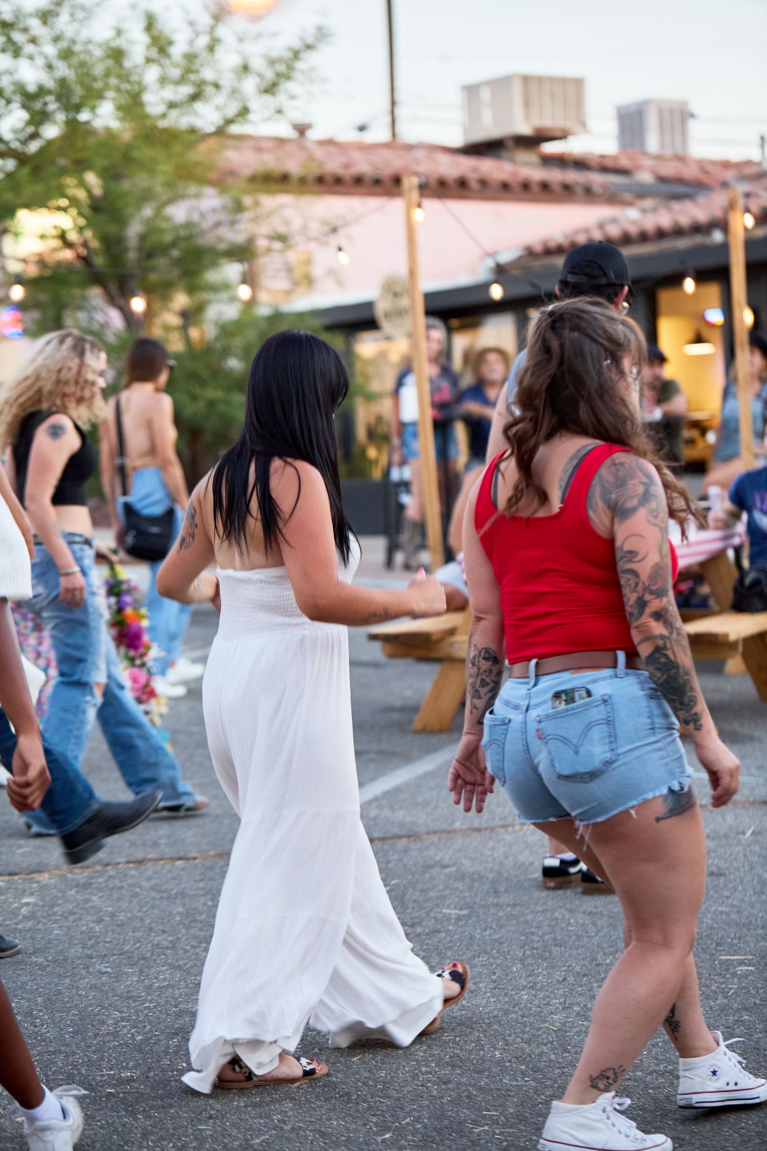 Two women dancing outdoors at a street party, one in a white dress and one in a red tank top with denim shorts and tattoos.