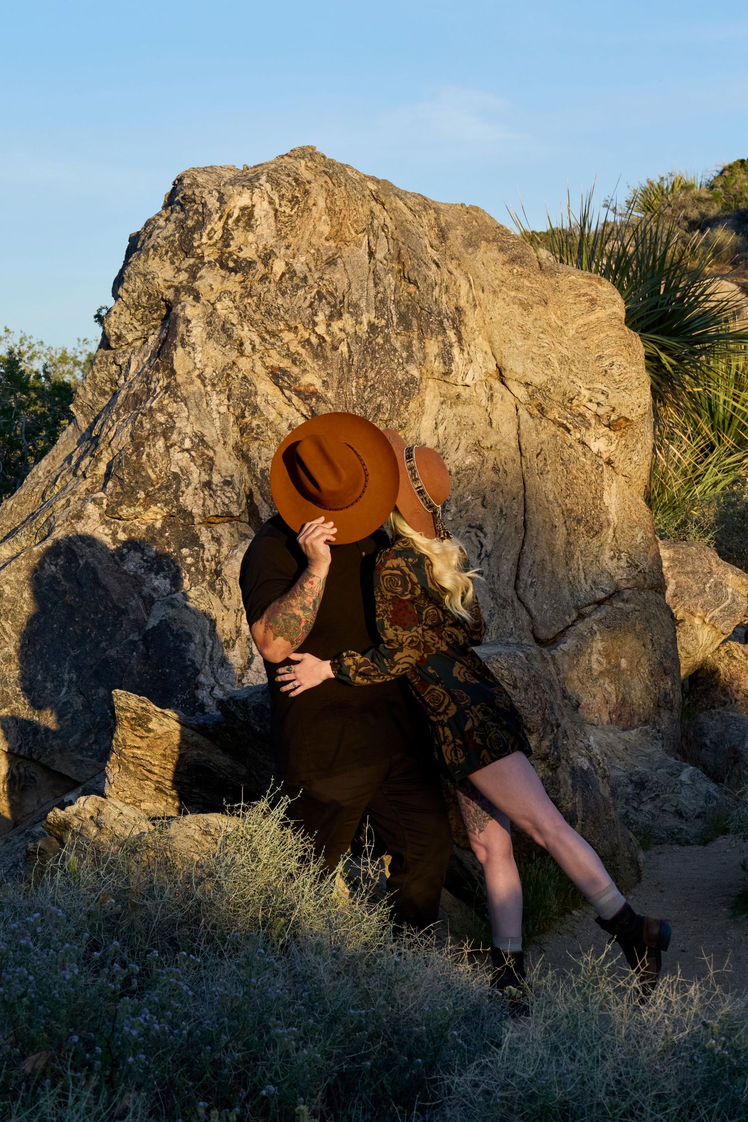 Couple kissing behind a brown cowboy hat against a large boulder in a desert landscape with blue sky.