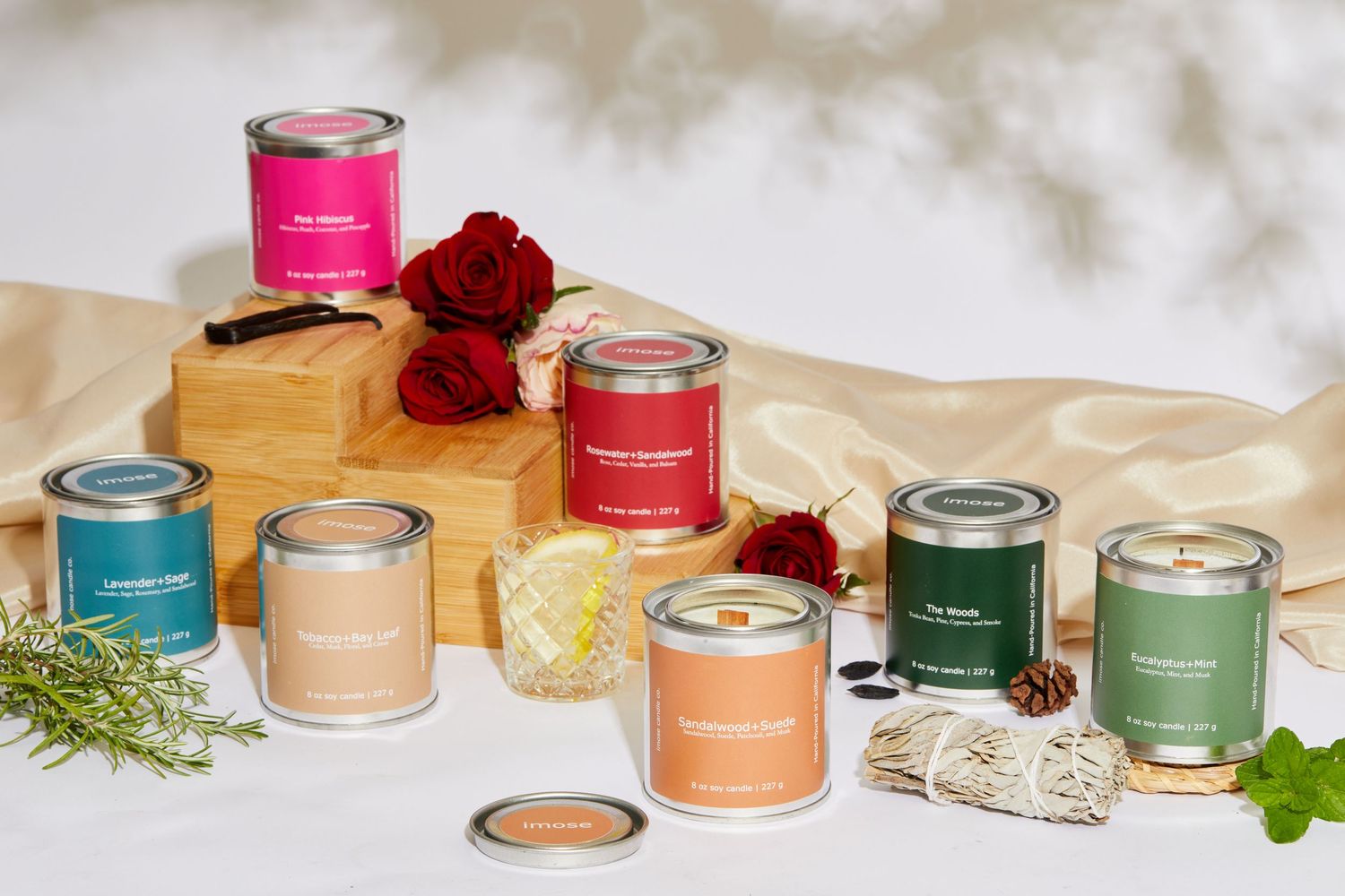 Collection of colorful scented candles arranged with red roses and herbs on a beige surface.