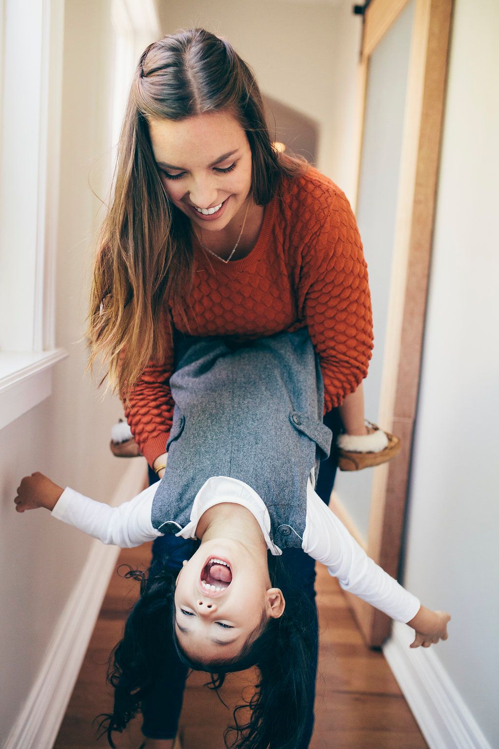 Playful parent holding child upside down in hallway with natural lighting.