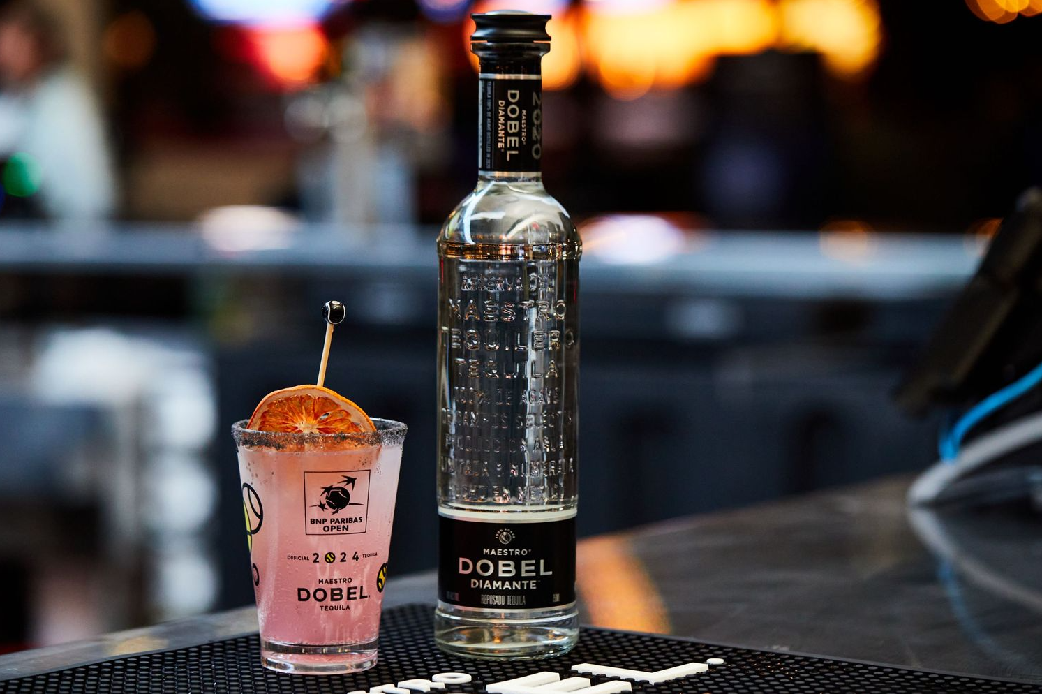 Glass bottle and pink cocktail photographed against blurred nighttime background.