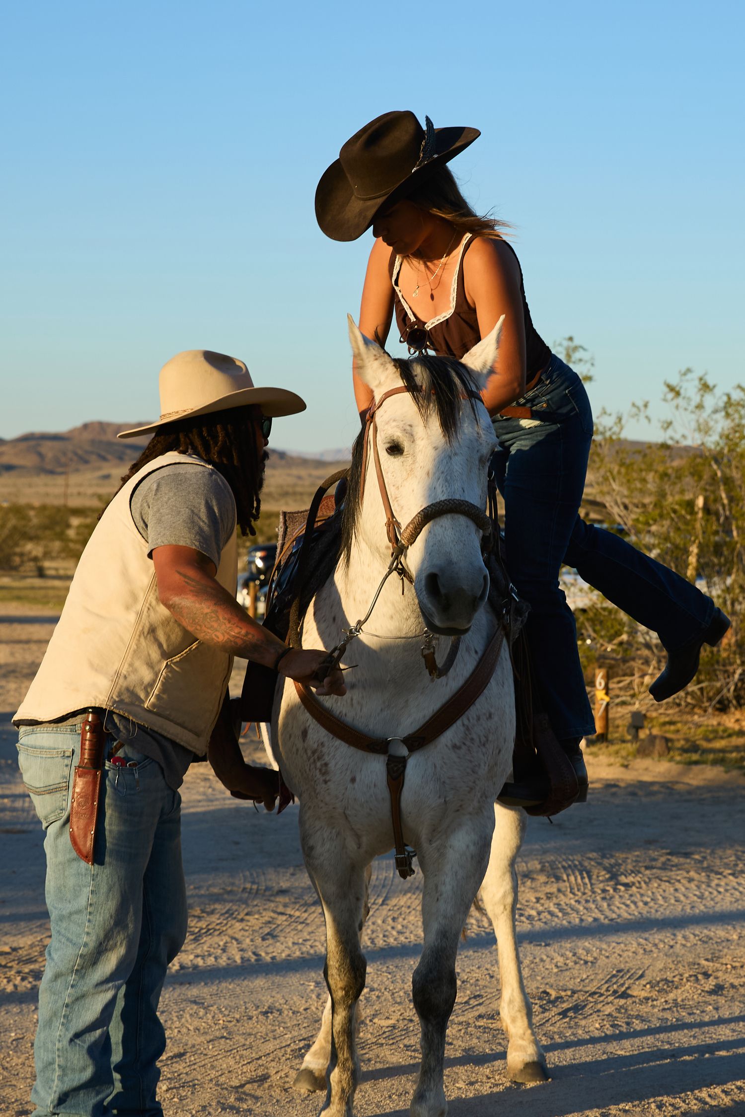 Cowboy helping a woman in a black hat mount a white horse at a sunny desert ranch.