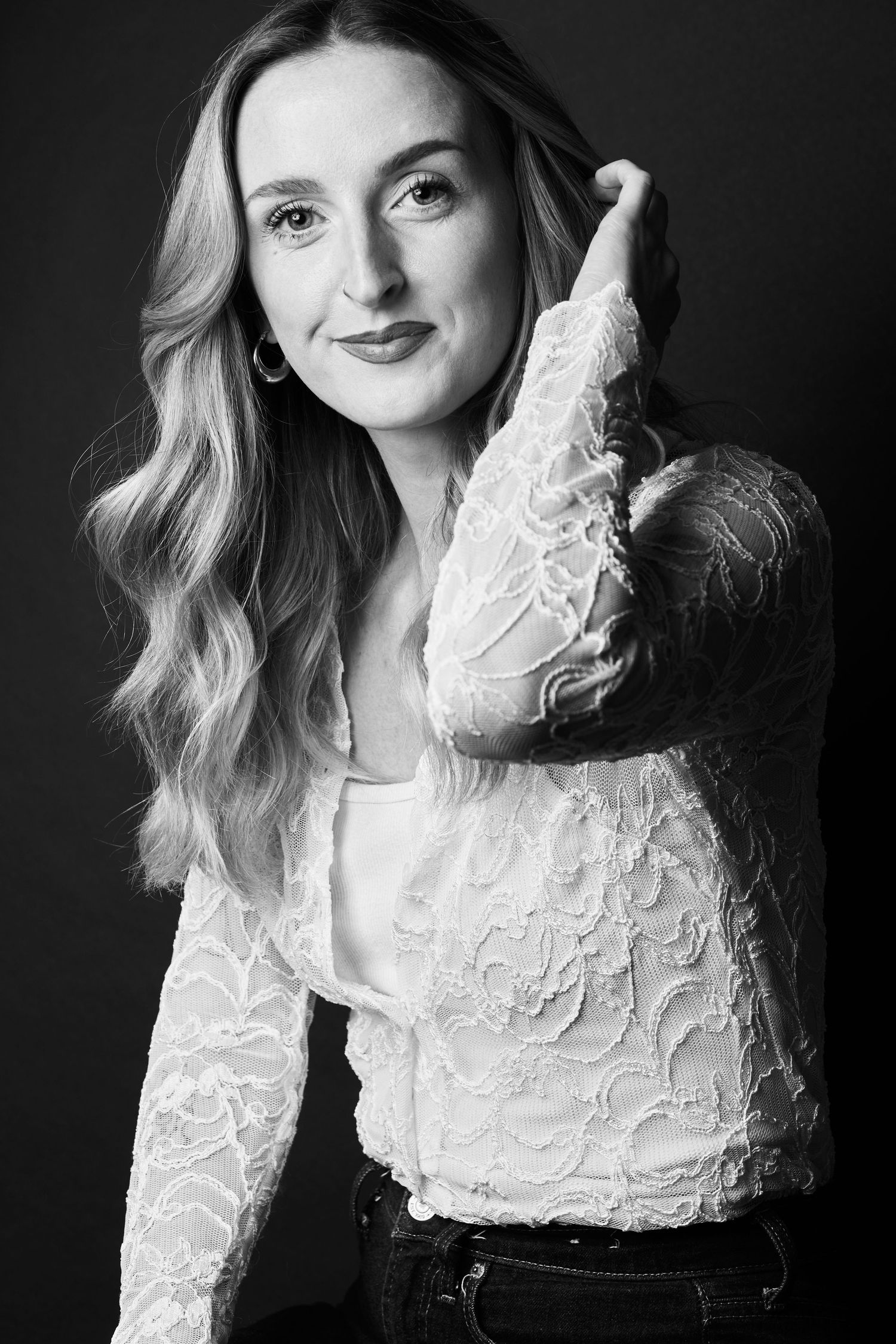 Black and white portrait of a woman in lace top, posing with hand raised, wavy hair, dark background.
