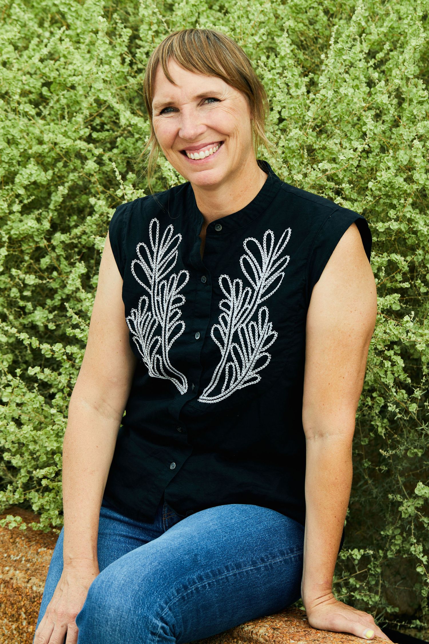Person wearing sleeveless black top with white leaf design poses outdoors against green foliage.