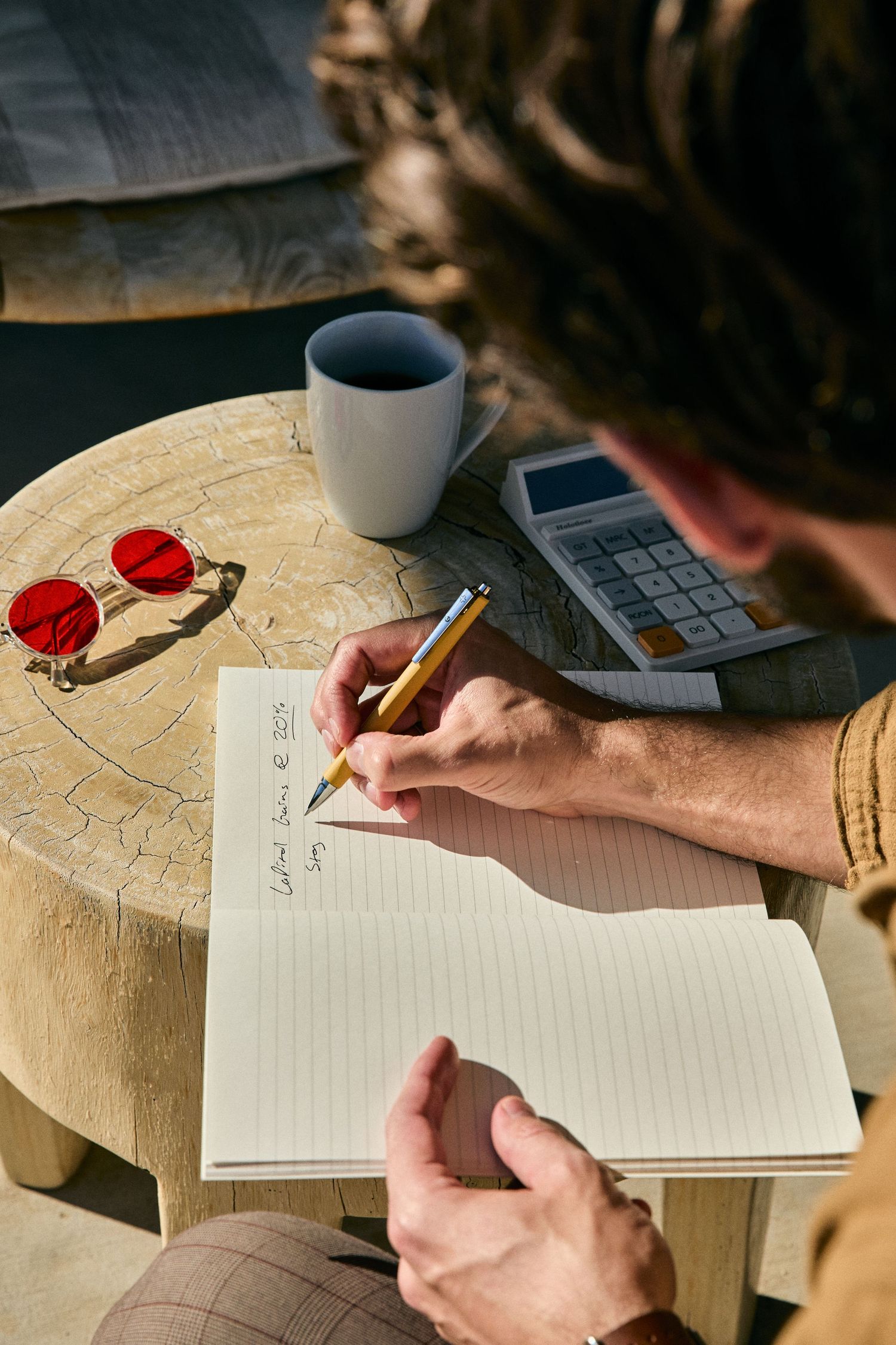 Person writing in a notebook with a cup of coffee on a wooden table while working on a laptop.