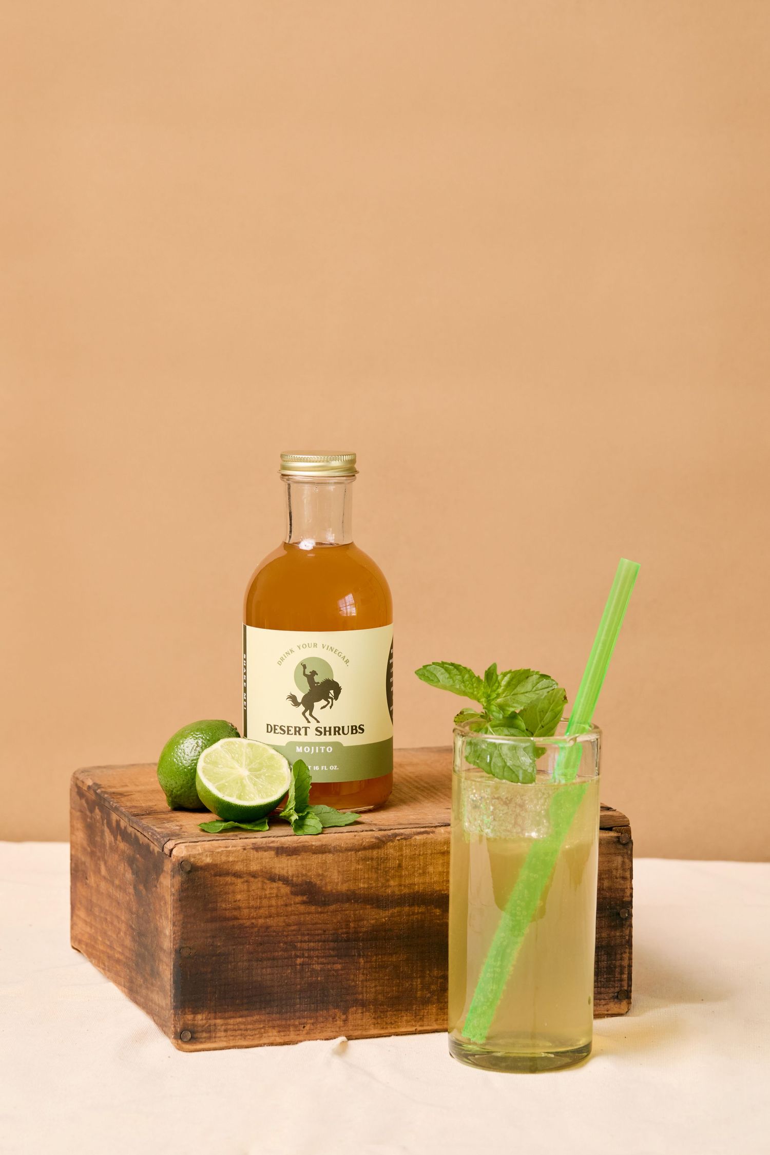 Desert Shrubs bottle with lime mojito cocktail in glass with green straw, fresh mint, and limes on rustic wooden box.