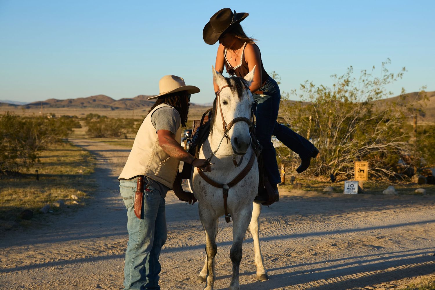 A woman in a cowboy hat rides a white horse while a man in a hat assists her in a desert landscape.