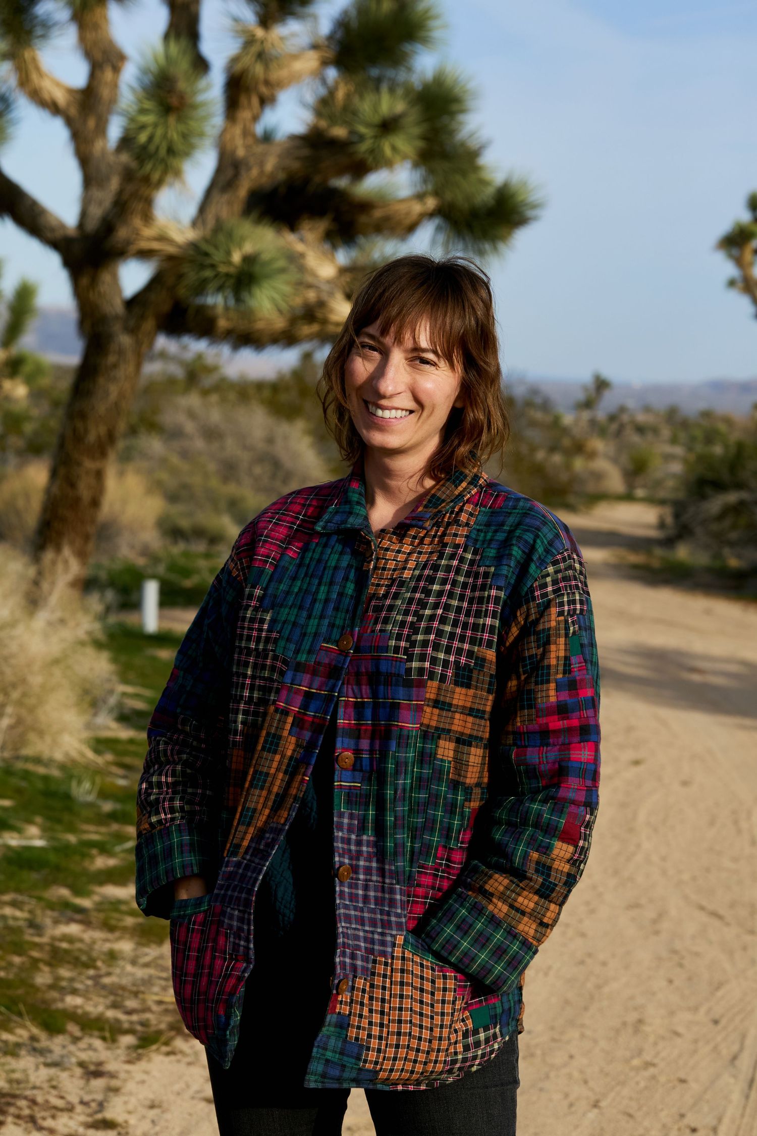 Woman smiling in colorful patchwork plaid jacket on desert dirt path with Joshua trees in background.