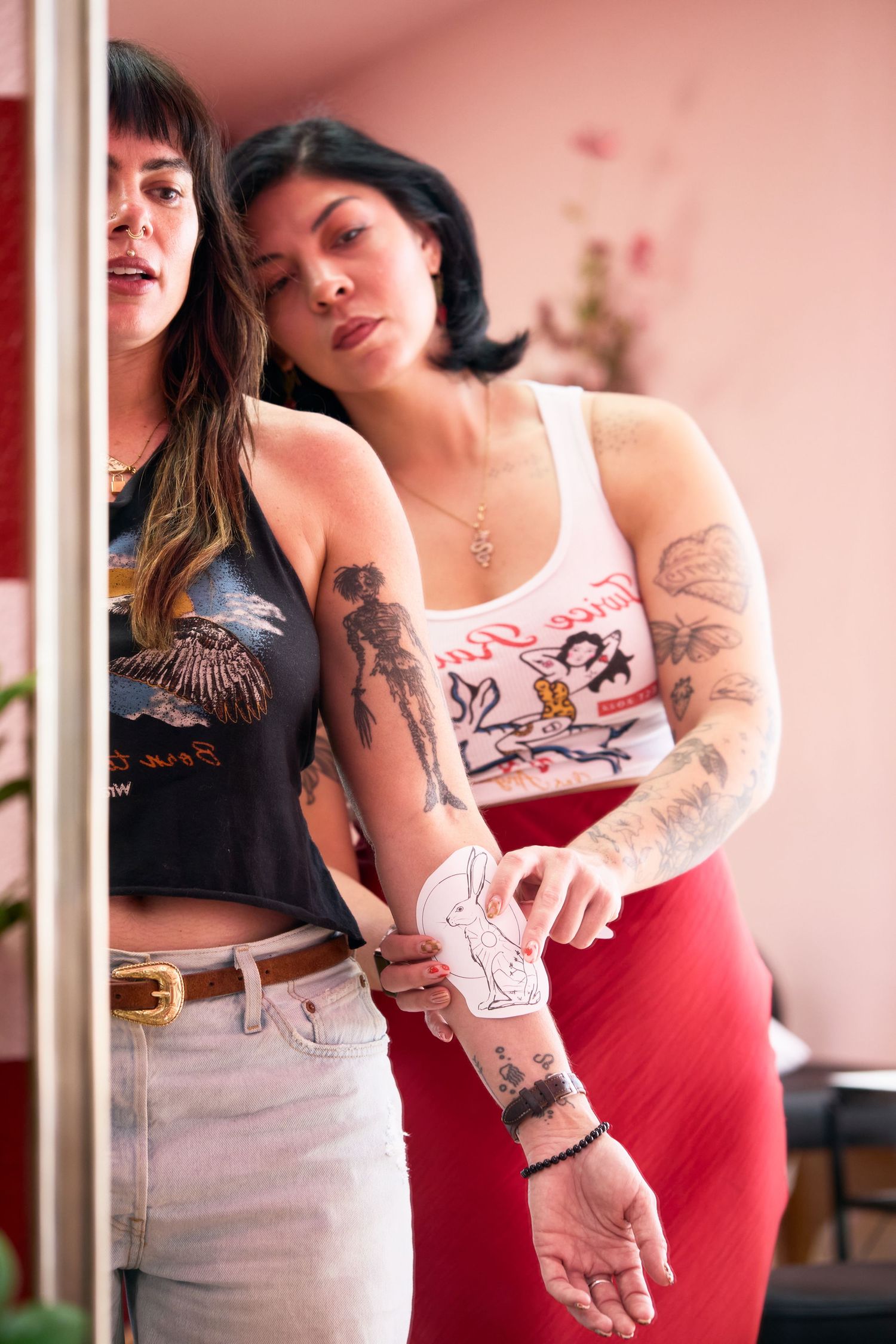 Two tattooed women pose together, one in a graphic crop top with jeans, the other in a white tank and red skirt.