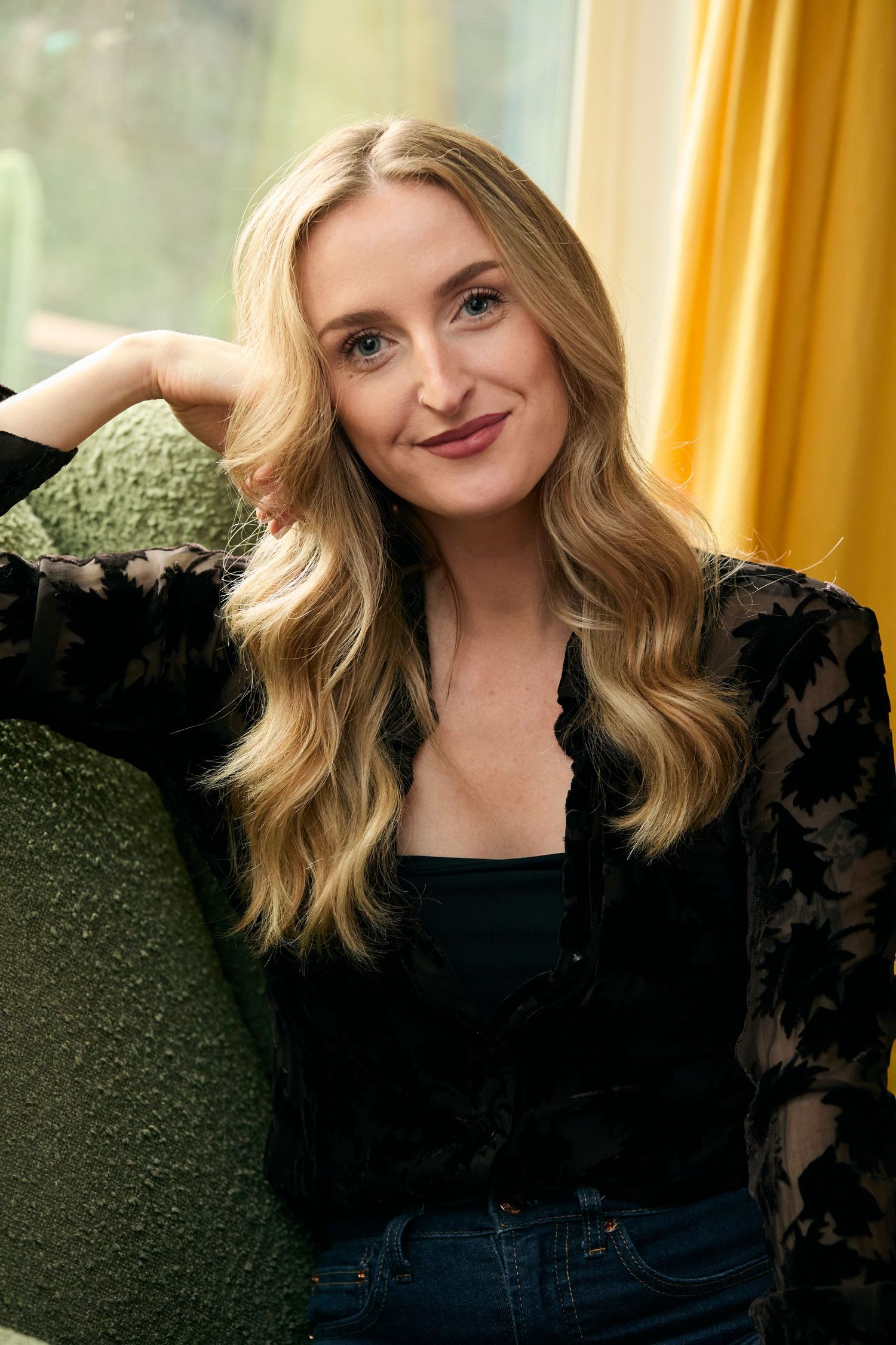 Blonde woman in black lace jacket smiling, seated against green chair with yellow curtain background.