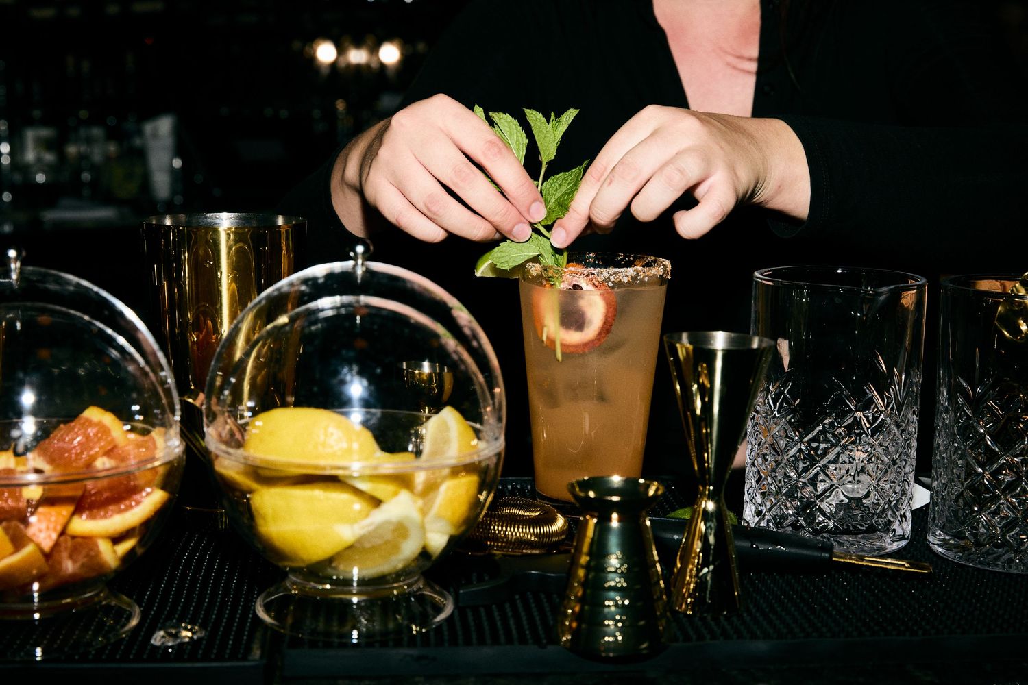 Bartender garnishing cocktails at a dark bar with various drinks and mixology tools on display.