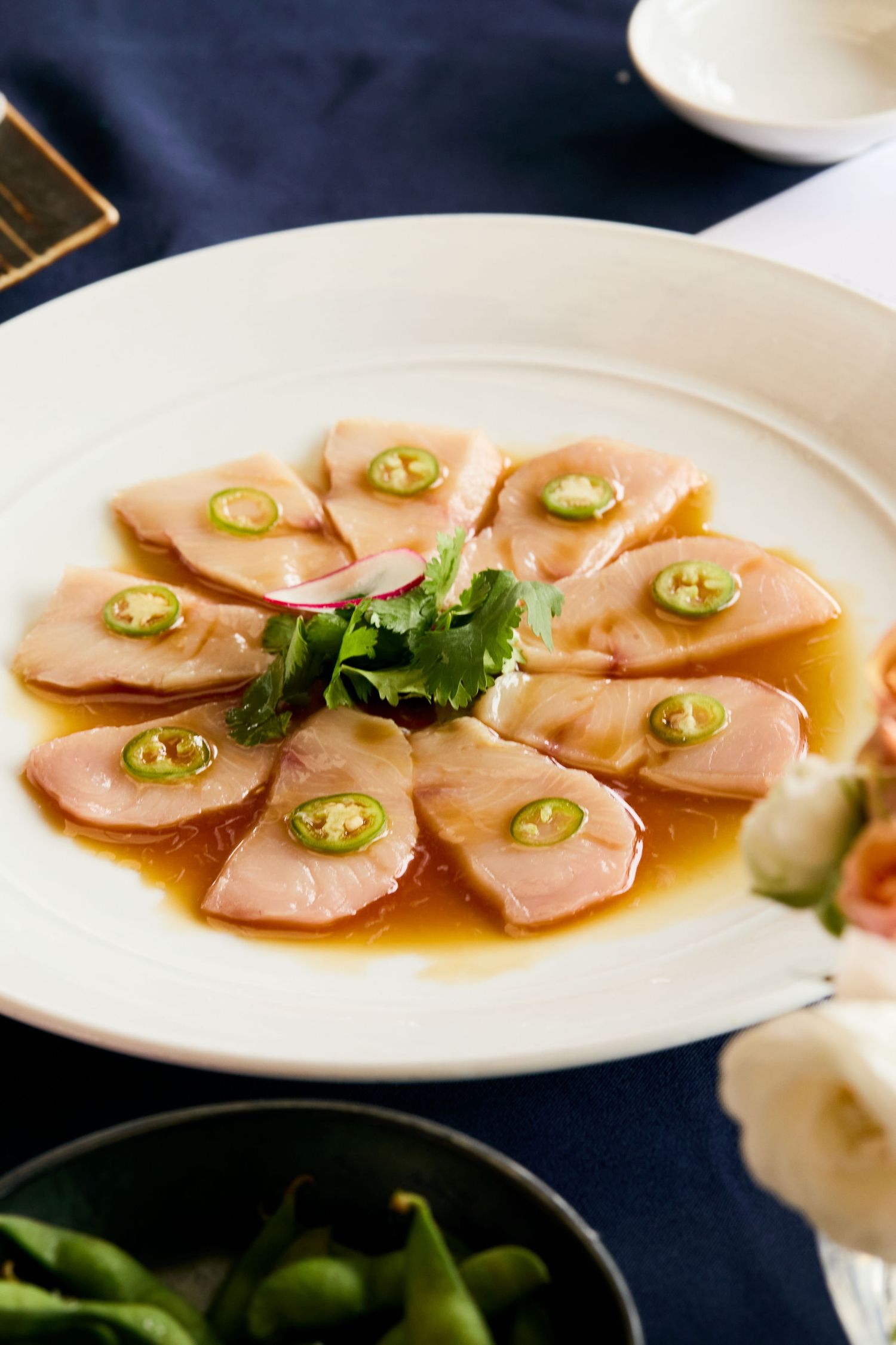 Thinly sliced yellowtail sashimi arranged in a circle with jalapenos and ponzu sauce on a white plate.