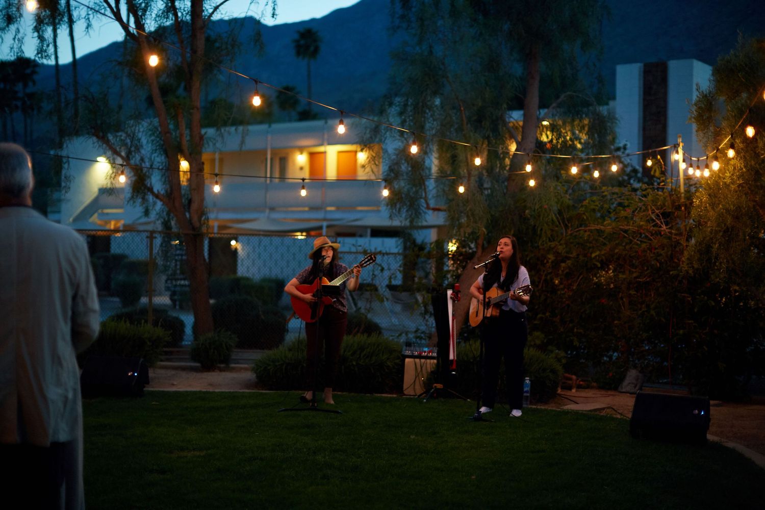 Musicians perform at an outdoor evening venue with string lights illuminating a garden setting at dusk.
