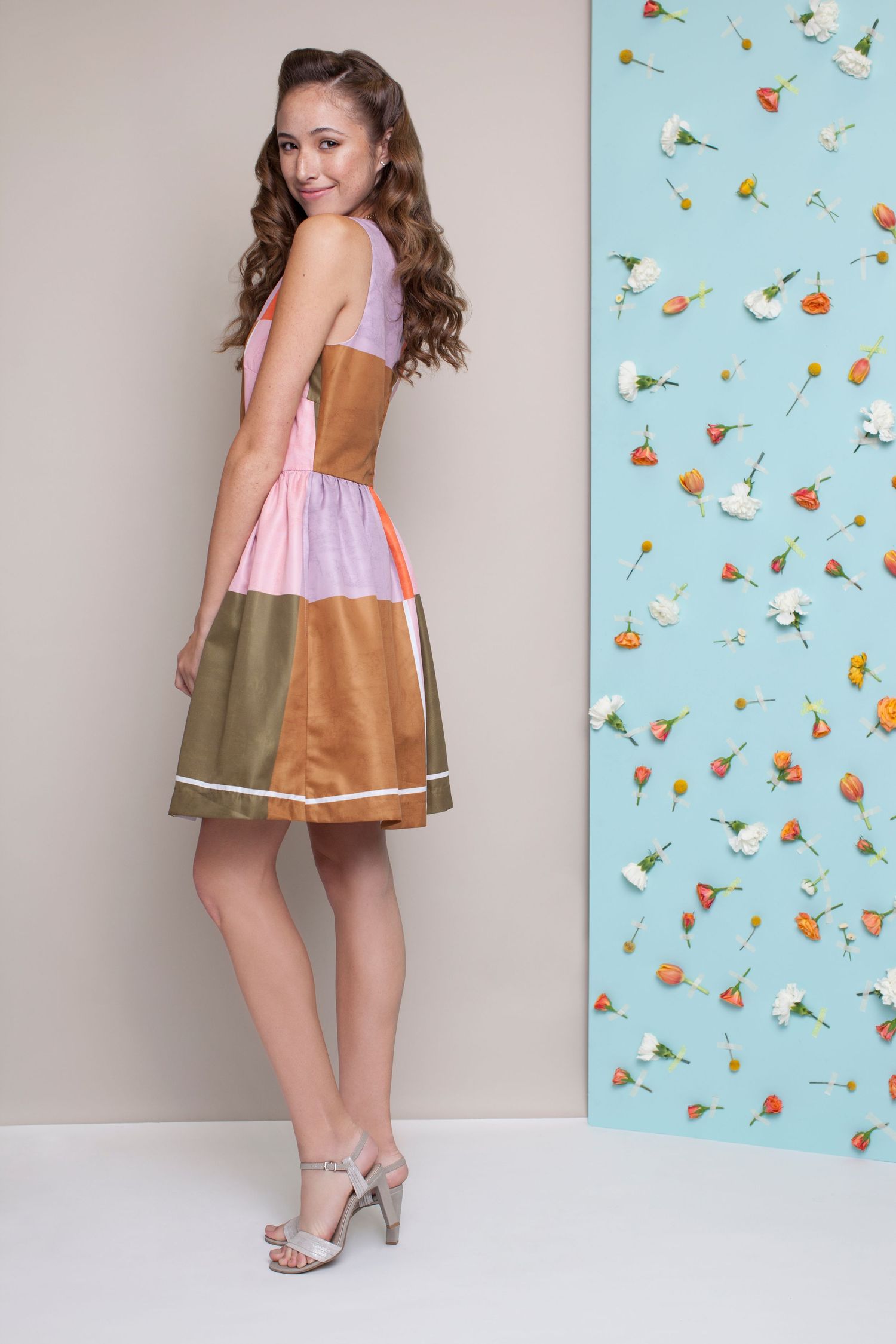 A model in a vintage-inspired colorblock dress with pink and brown tones poses next to floral wallpaper.