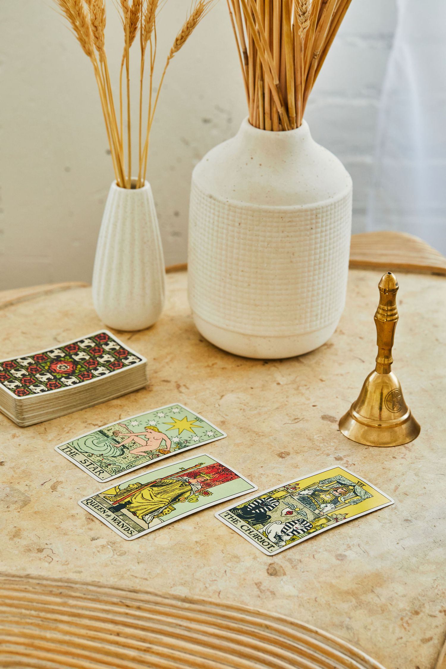 Tarot cards spread on a rustic wooden table with white ceramic vases holding dried wheat stems and a brass bell.