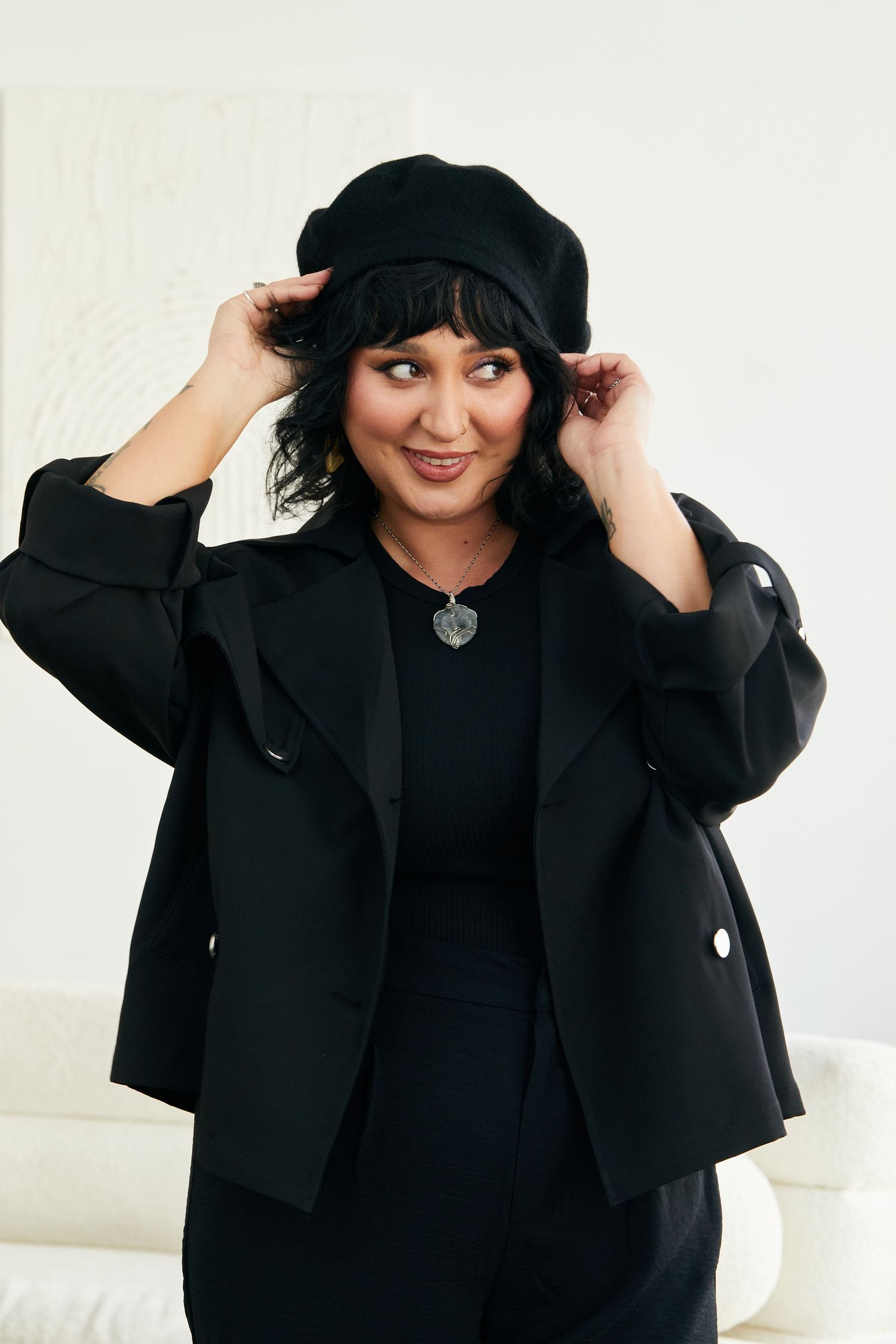 Woman in all-black outfit wearing beret hat and blazer, smiling in bright white room.