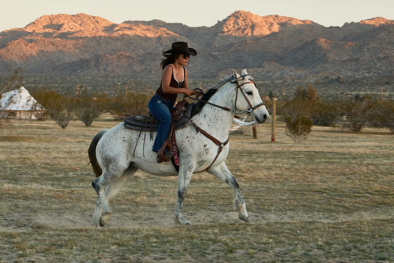 A woman in a black cowboy hat rides a white horse across a desert landscape with mountains at sunset.