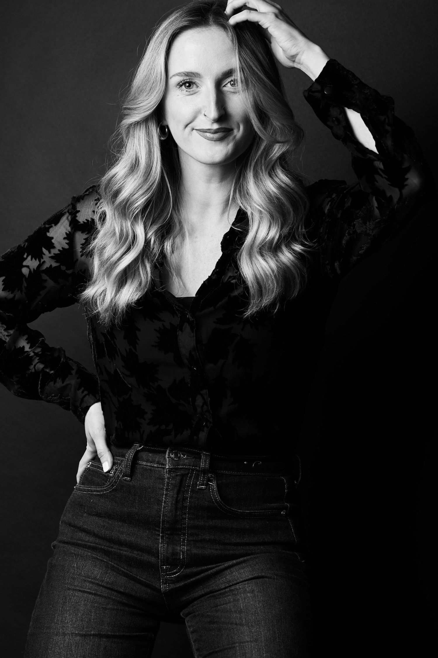 Black and white portrait of a blonde woman in lace blouse and jeans posing confidently against dark background.