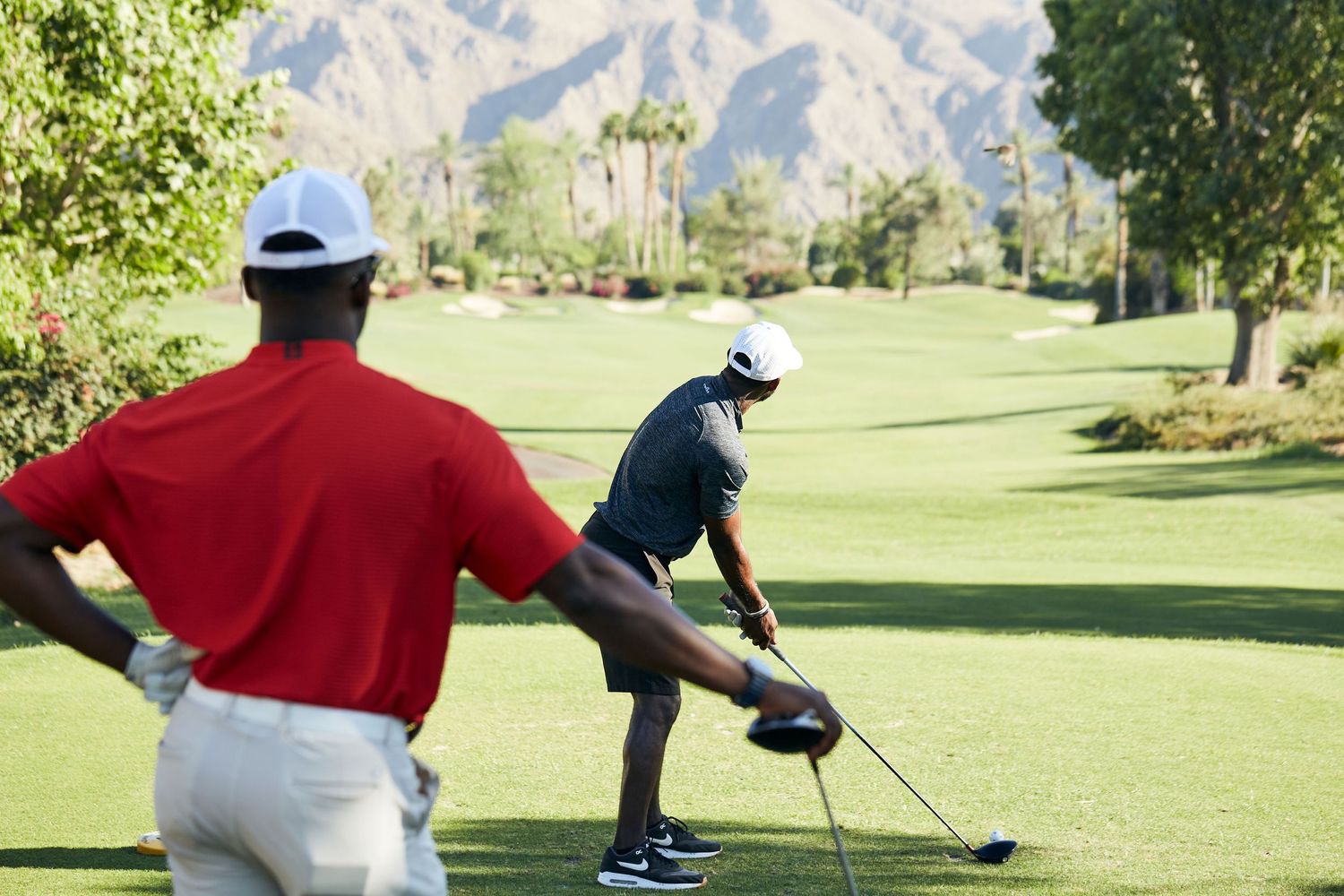 Two golfers practicing their putting form on a sunny golf course with mountains in the distance.