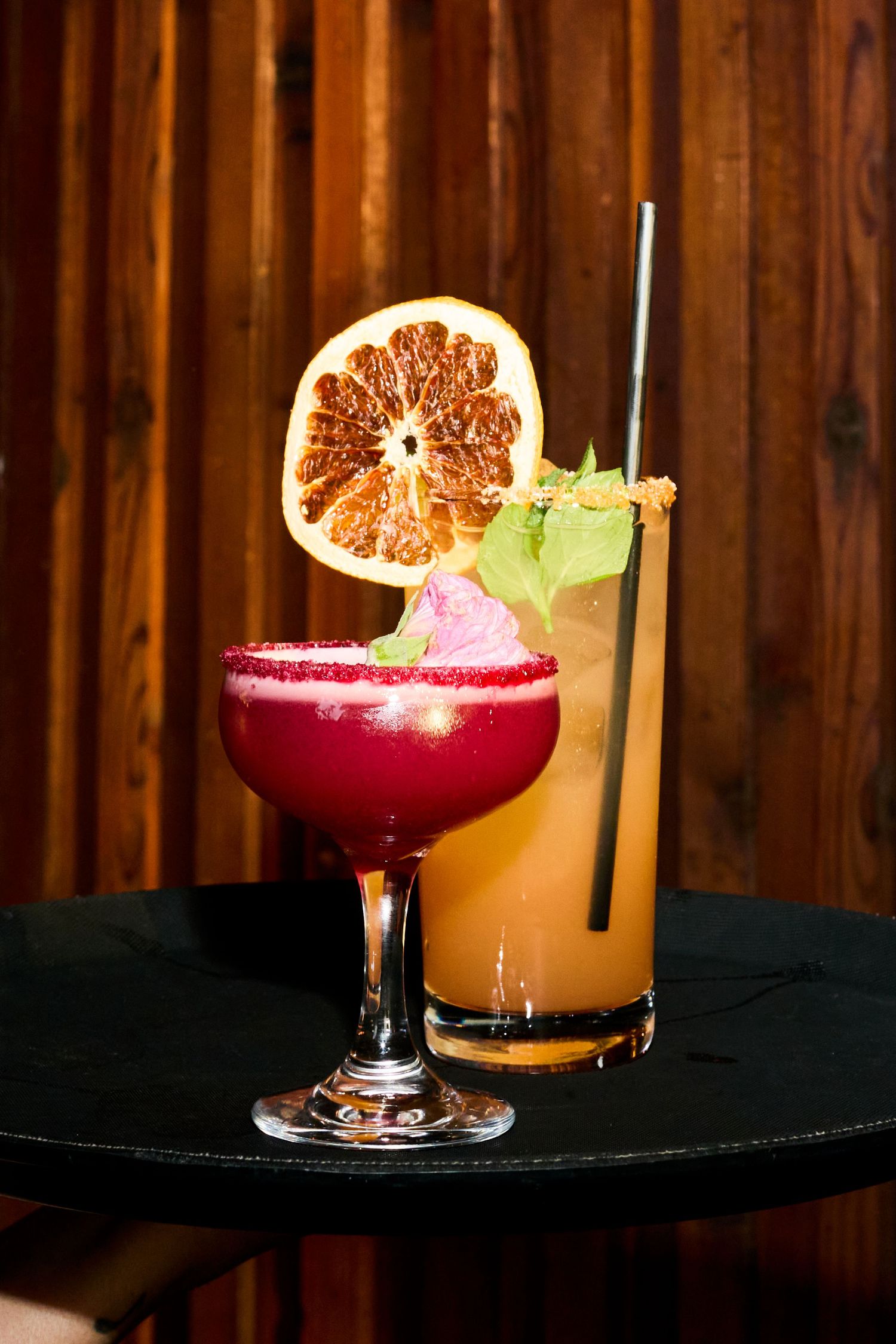 Two elegant cocktails on a black table, one red and one orange, garnished with citrus and herbs against a wooden backdrop.