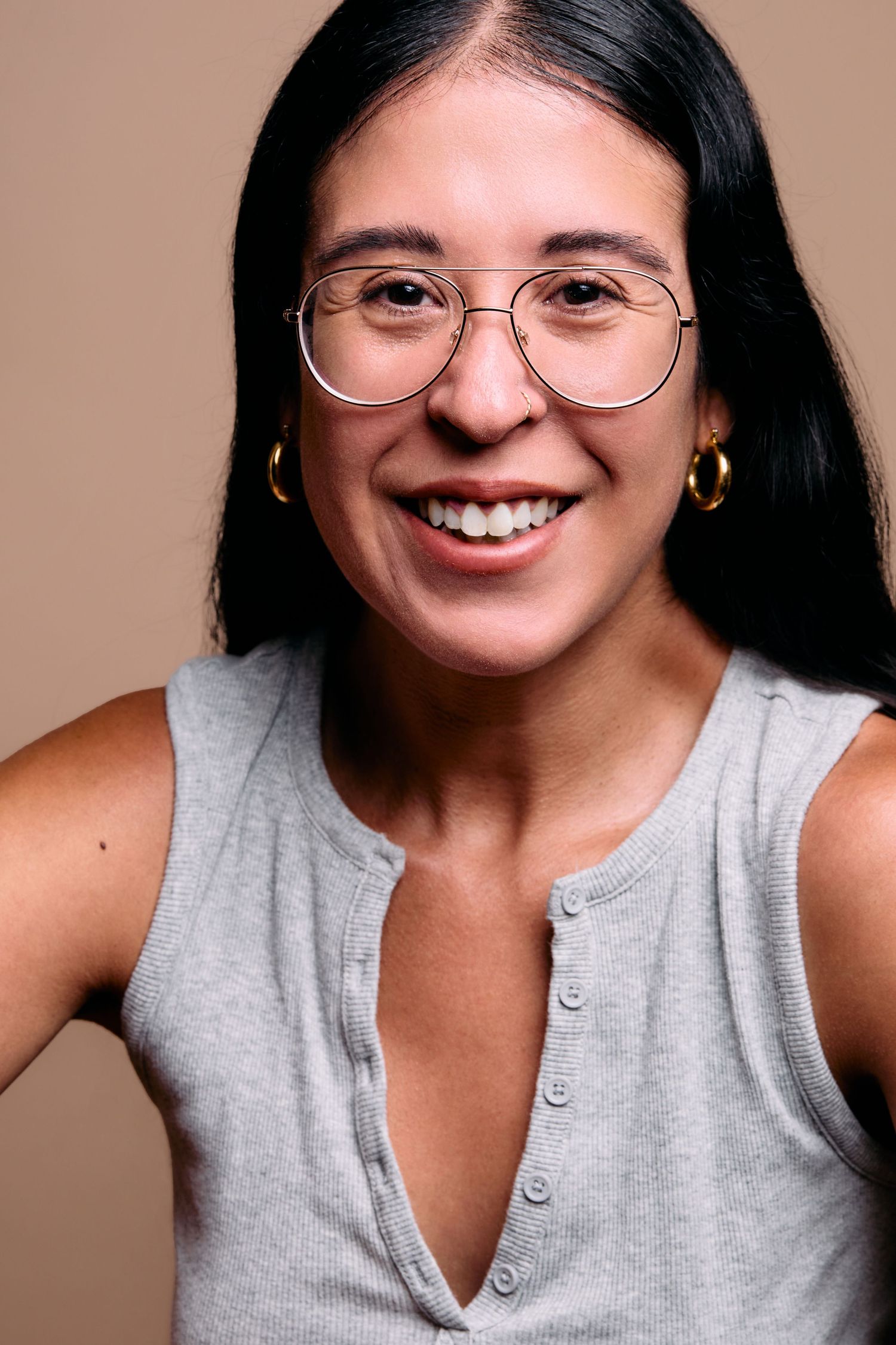 A person in a grey sleeveless top and circular glasses smiles warmly against a neutral beige background.