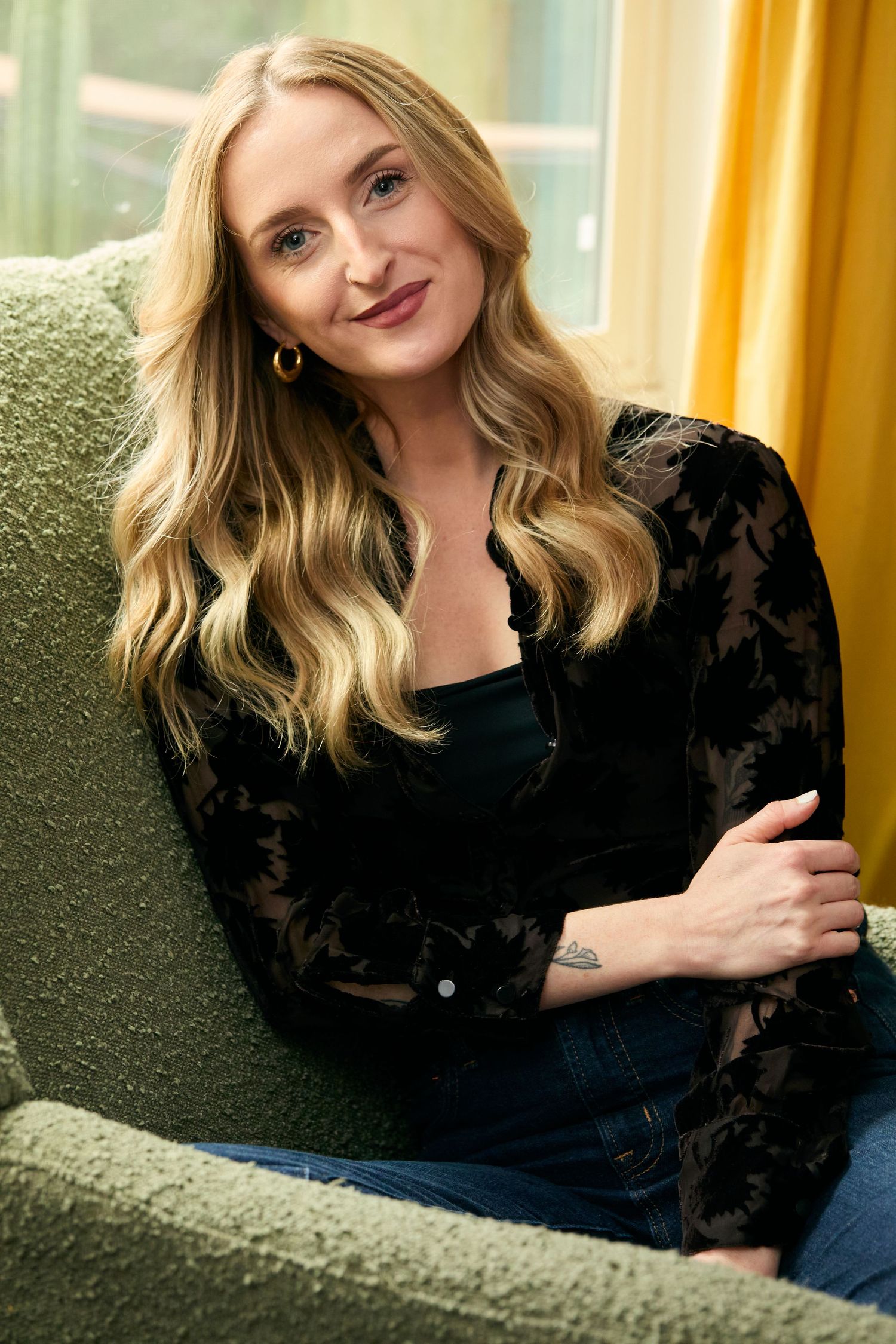 Smiling blonde woman in black lace top and jeans sitting relaxed on green chair near yellow curtains.