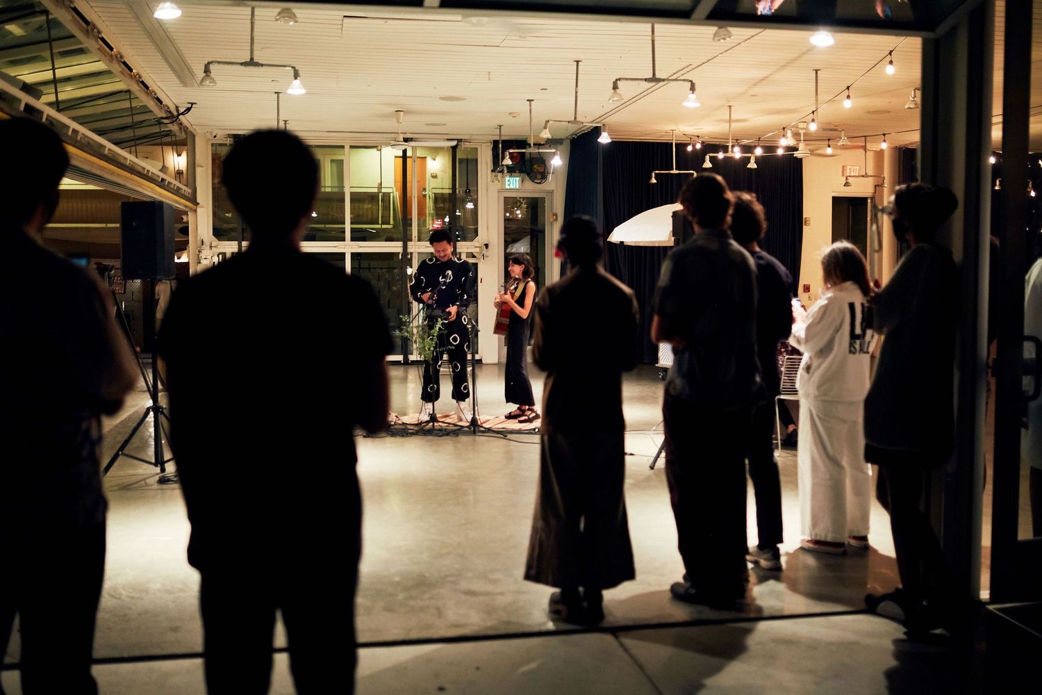 People gather in silhouette at a dimly lit indoor venue with warm lighting and reflective surfaces.