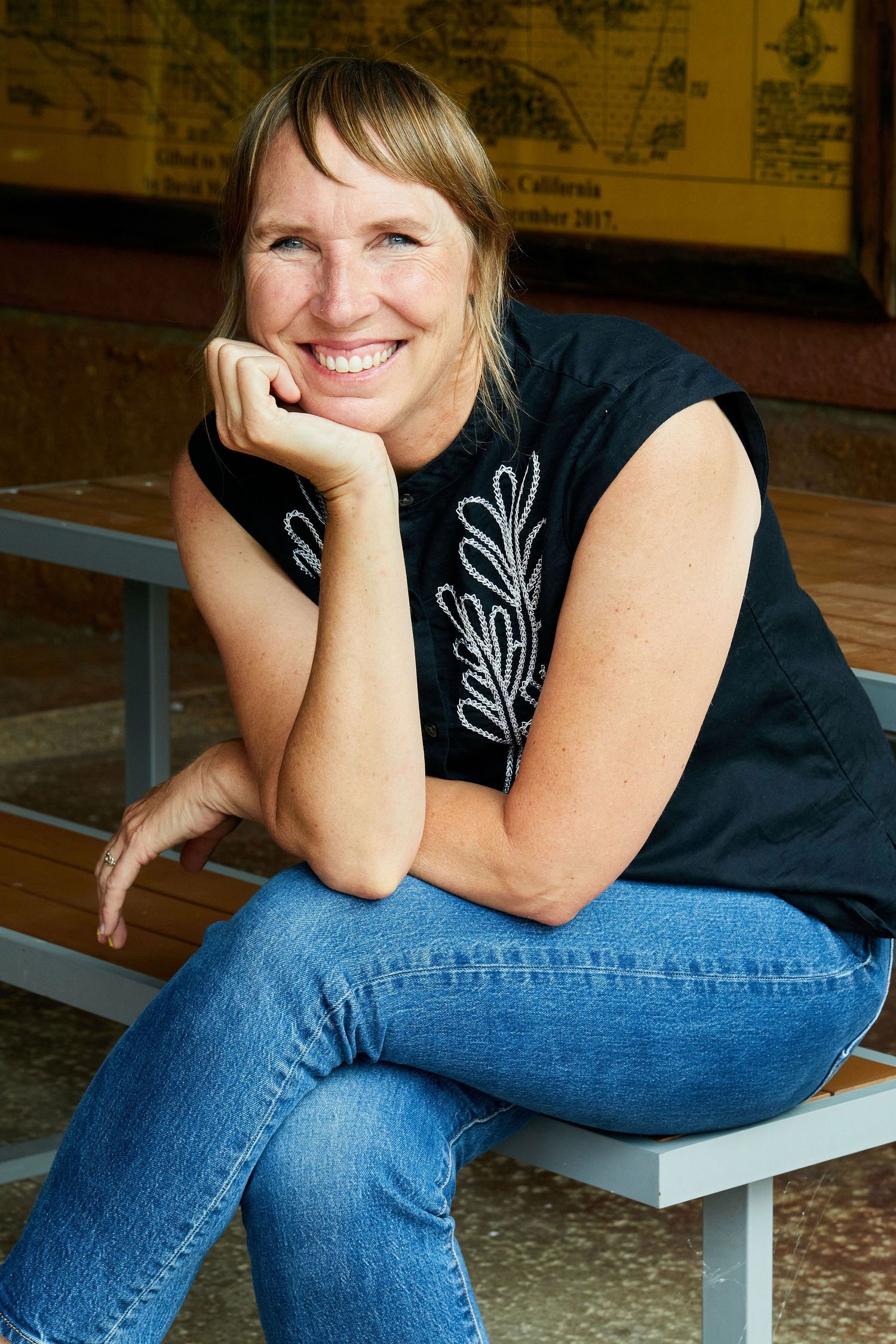 Person in black sleeveless top with white embroidery sits casually on bench smiling warmly.