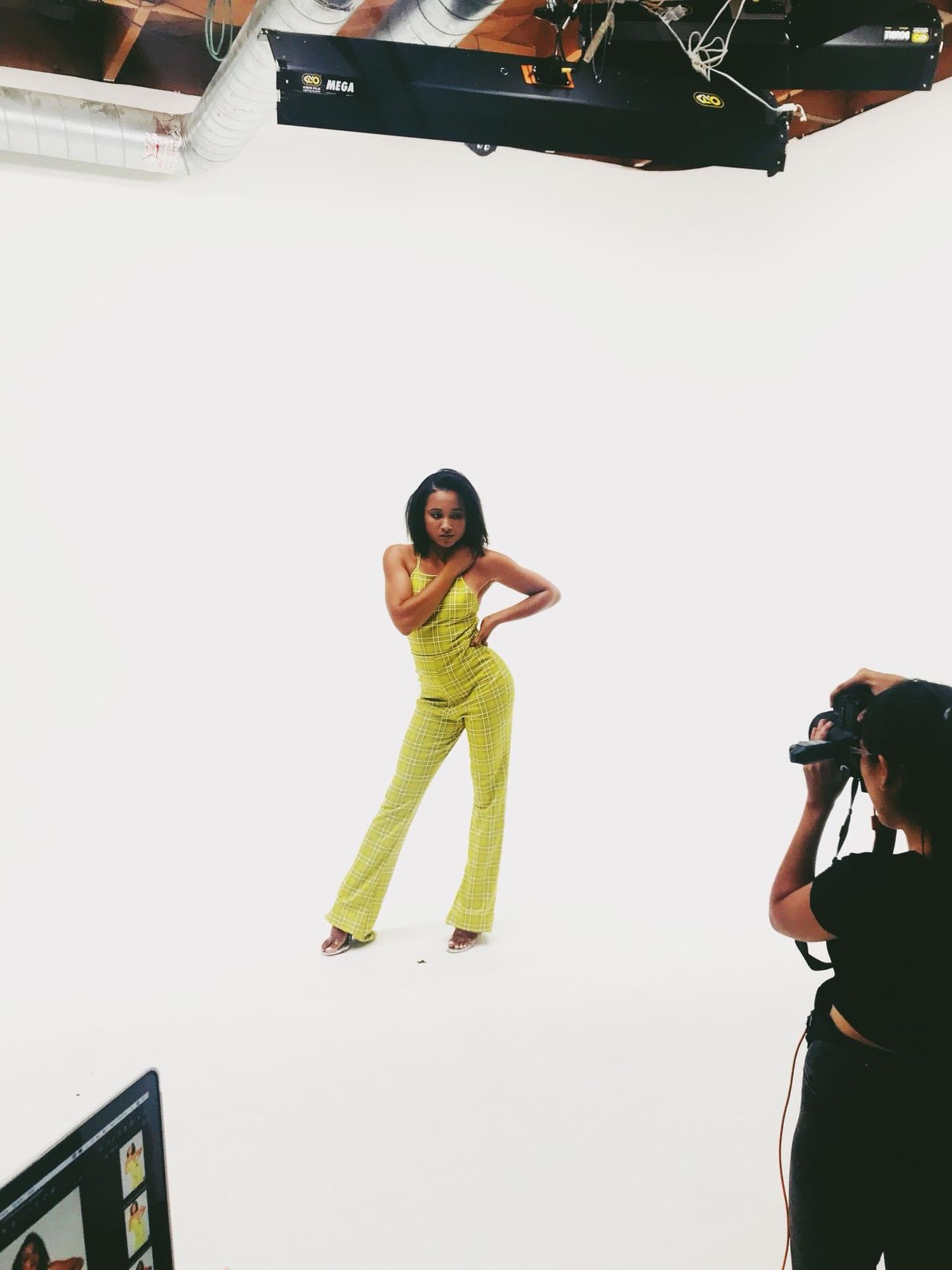 Behind-the-scenes fashion photoshoot showing a model in bright lime green jumpsuit posing against white backdrop with photographer.