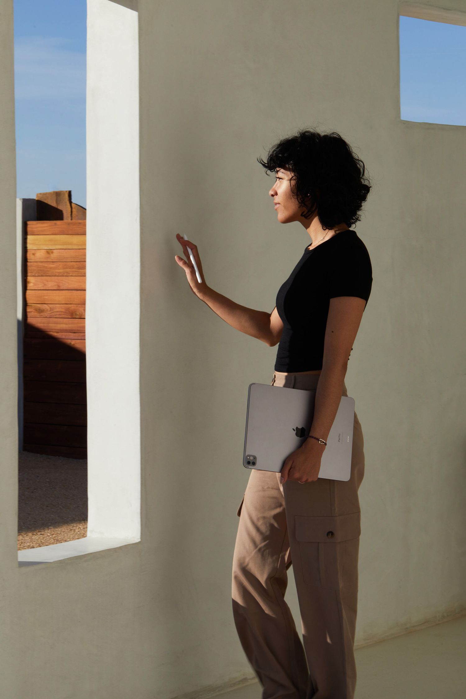 A person in a black top and khaki pants stands in profile against a white wall, holding a laptop while gesturing.