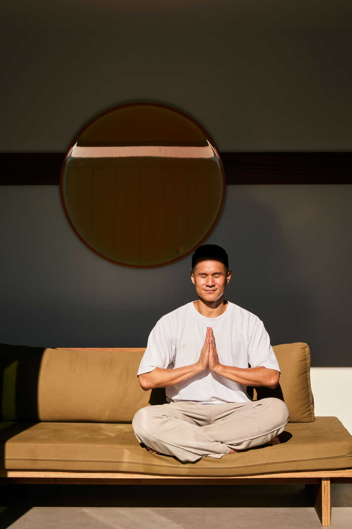 Person in white clothing sits cross-legged in meditation pose on tan couch beneath circular wall mirror.
