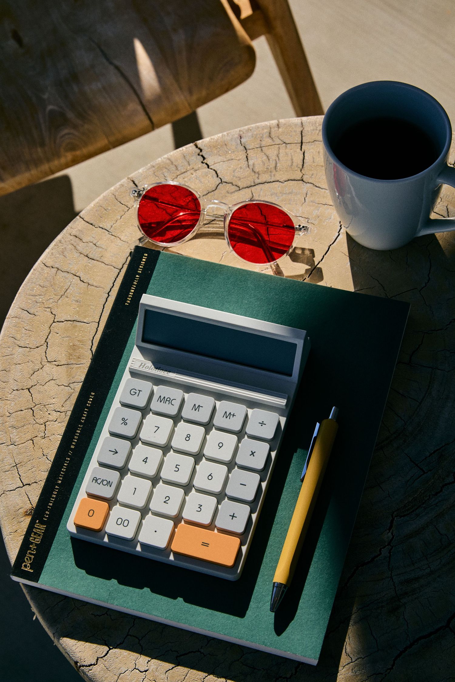 Retro calculator, red round sunglasses, and coffee mug on textured surface with green notebook and pencil in sunlight.