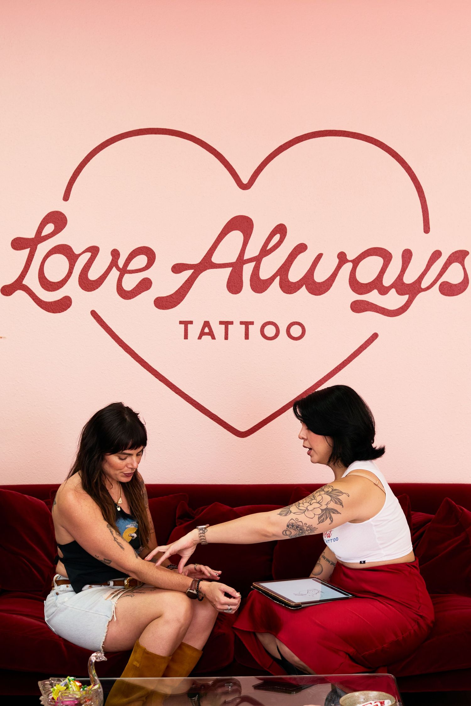 Two women consult at Love Always Tattoo studio, sitting on a red velvet sofa beneath a pink heart logo.