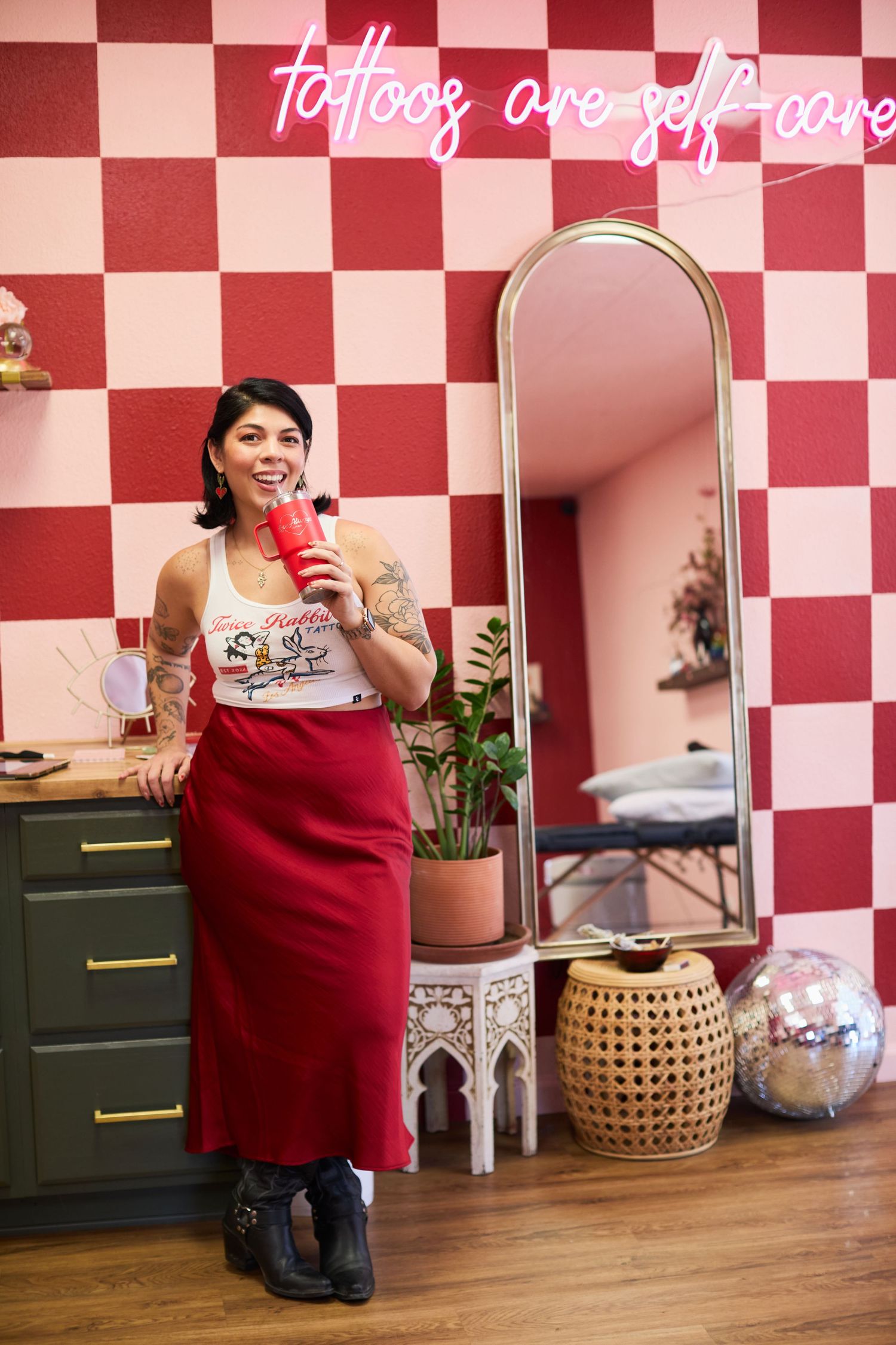 Woman in red skirt and tattooed arms holds red cup in colorful tattoo studio with neon sign reading tattoos are self-care.