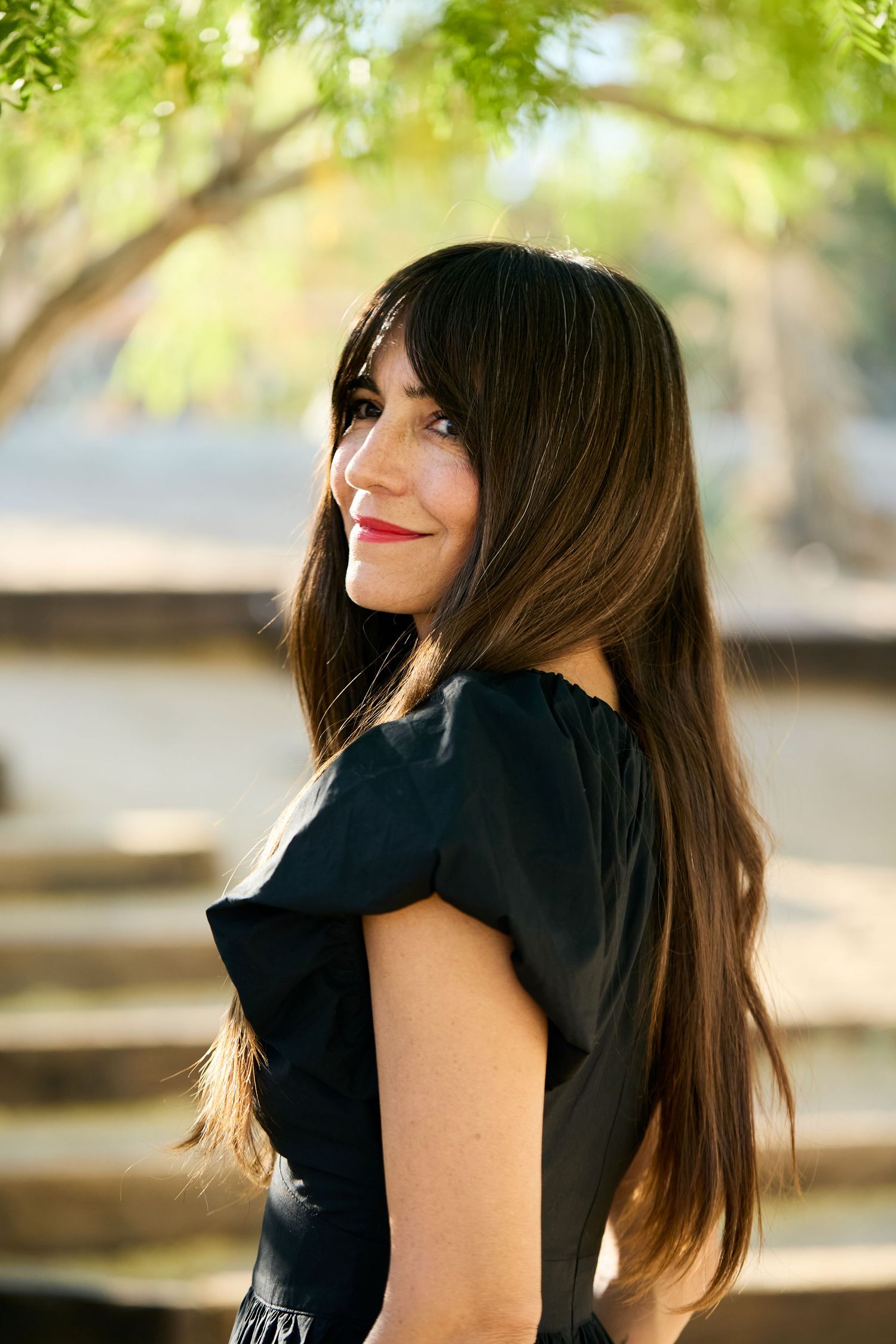 Woman in black dress poses outdoors with blurred green foliage background in natural lighting.