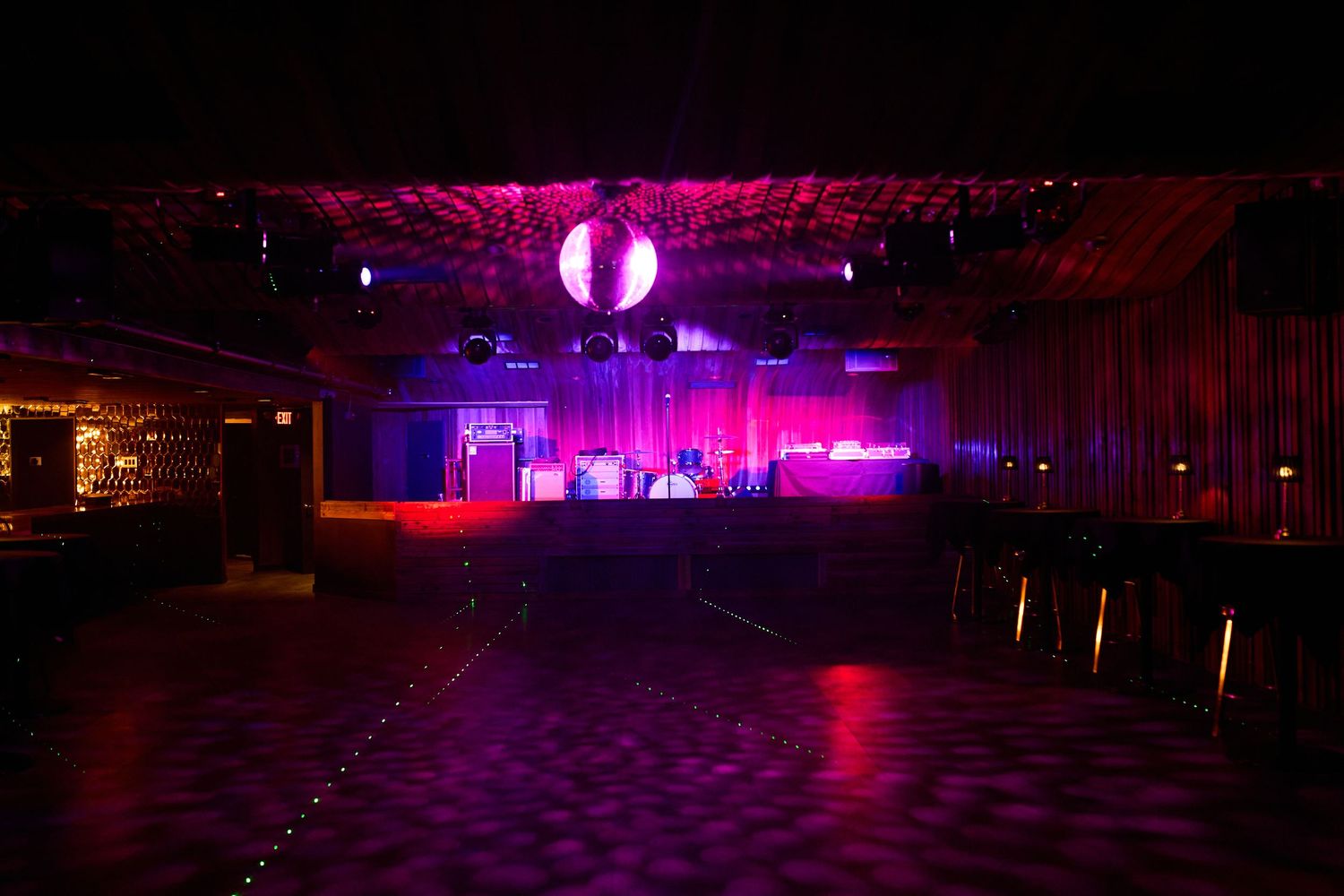 A dark nightclub interior with purple and pink stage lighting illuminating an empty dance floor and performance stage.