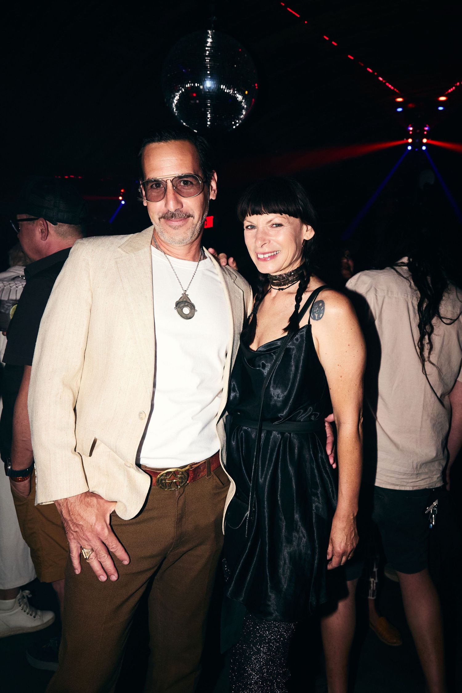 Two people pose together at a nightclub with disco lighting, one wearing a light blazer and one in a black dress.