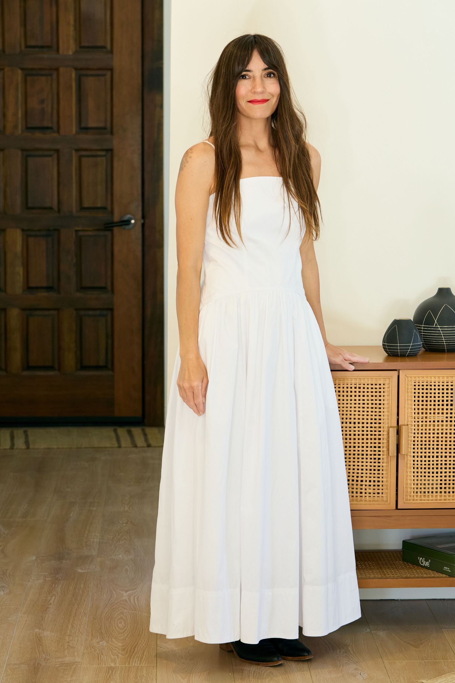 Woman in elegant white strapless maxi dress stands beside wooden cabinet on hardwood floor.