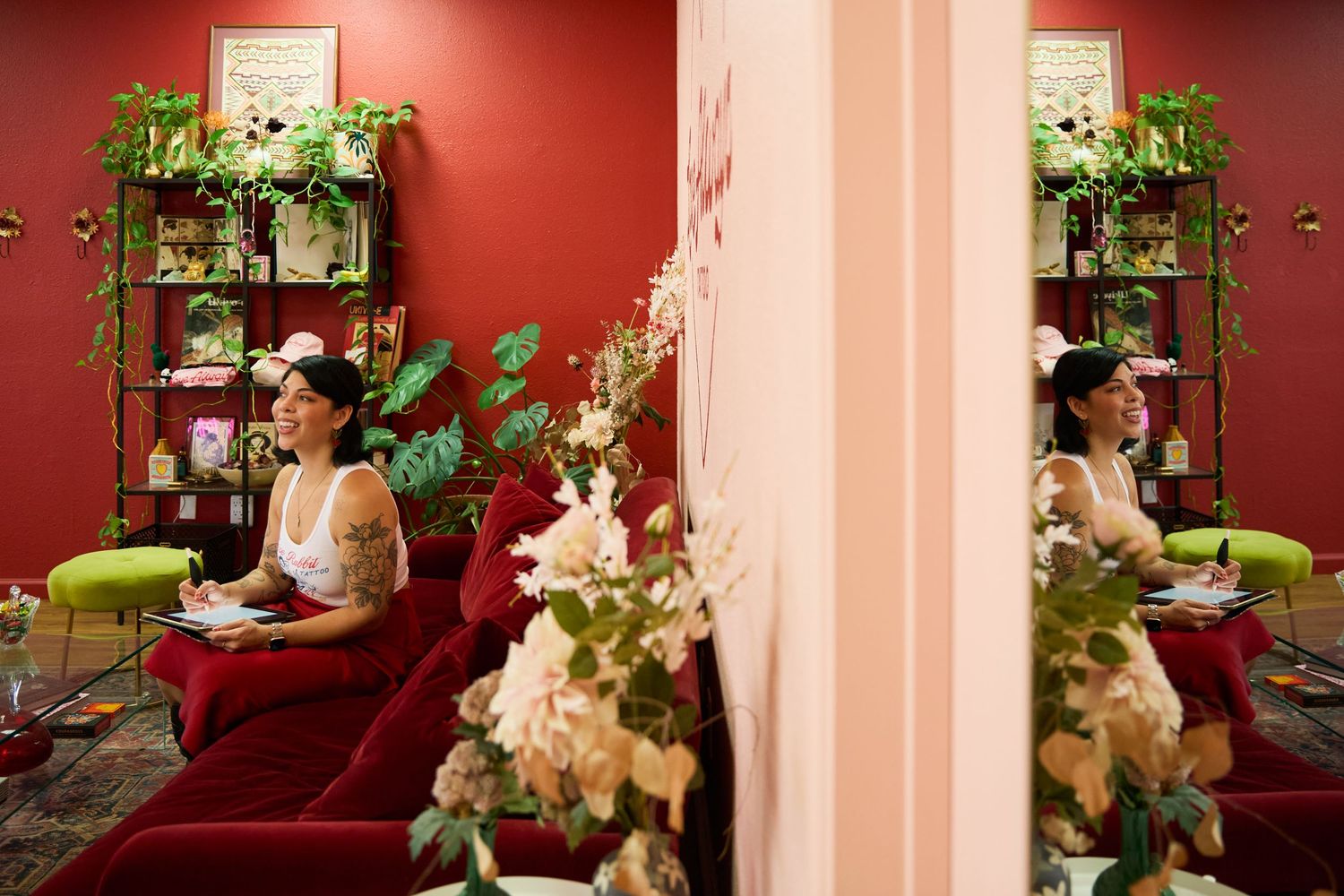 Two tattooed women sit on red velvet sofas in a lush, plant-filled salon with warm red walls and floral decor.