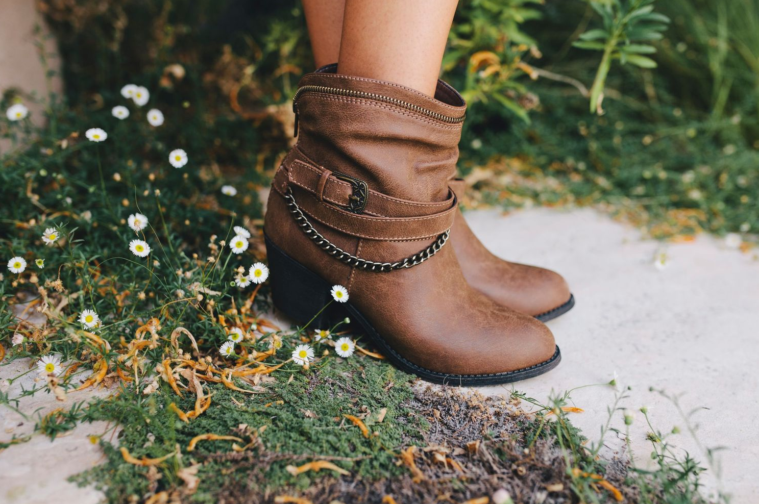 Brown leather ankle boots with chain detail shown against small white flowers and grass.