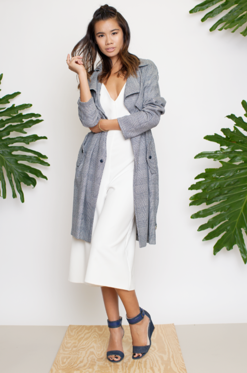 Fashion model wearing grey coat over white dress with tropical leaves backdrop.