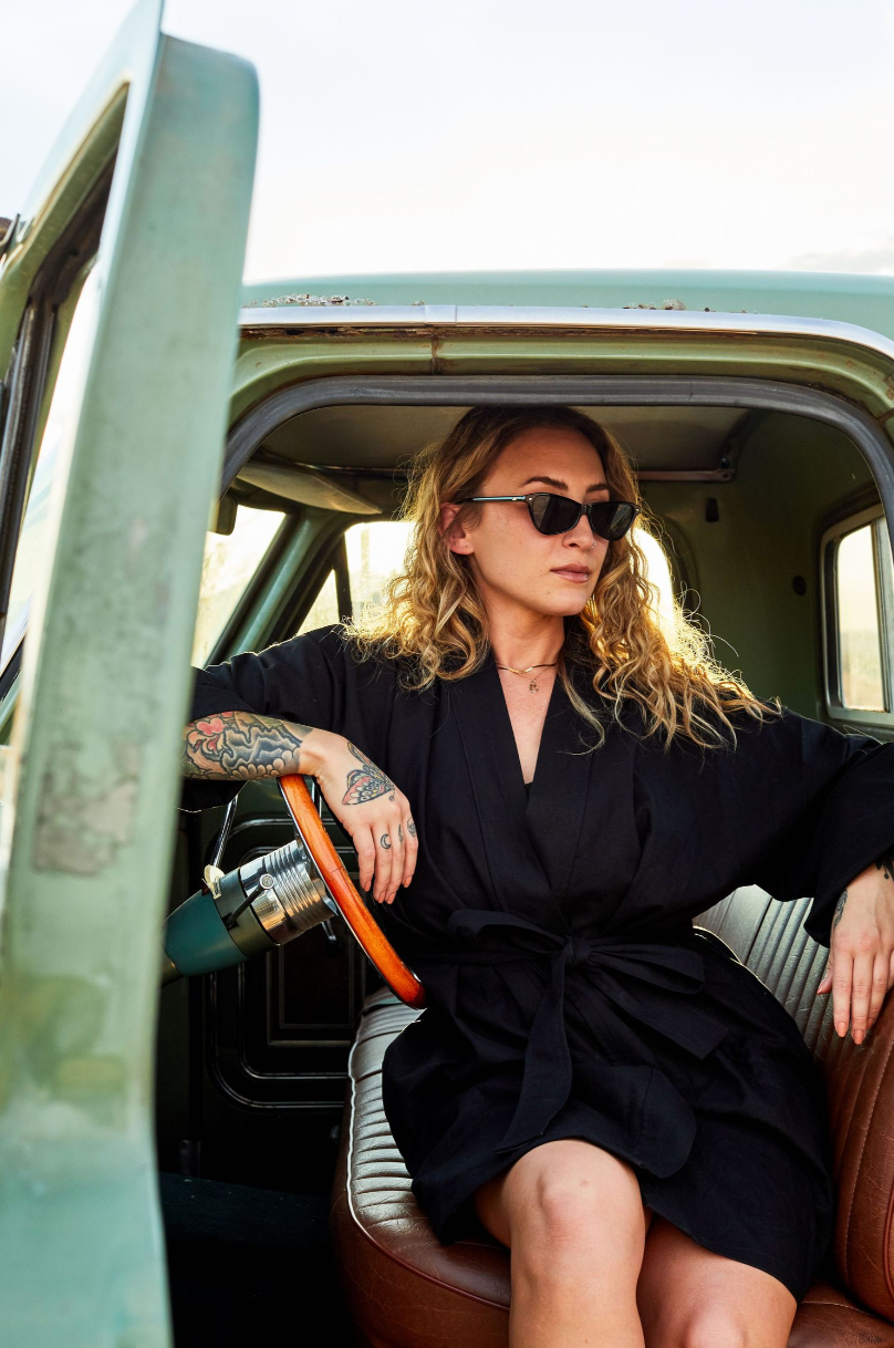 Stylish portrait in vintage car showing someone in black dress and sunglasses with tattoos.