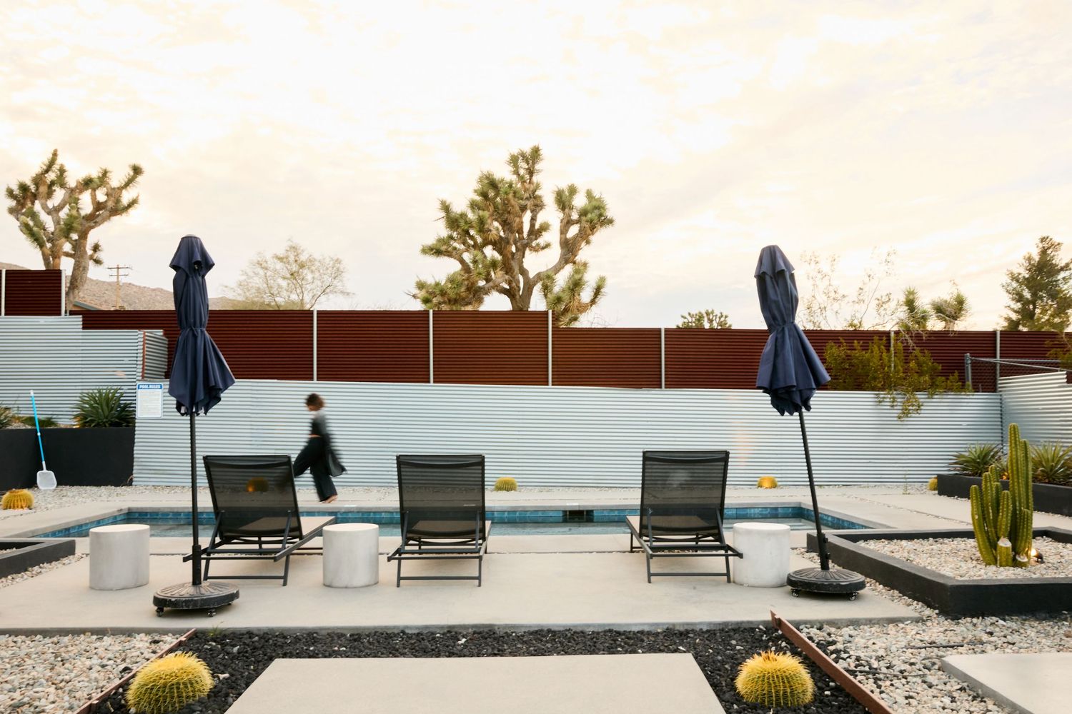 Modern poolside lounge area with three chaise lounges and navy umbrellas arranged on white gravel against a minimalist wall.